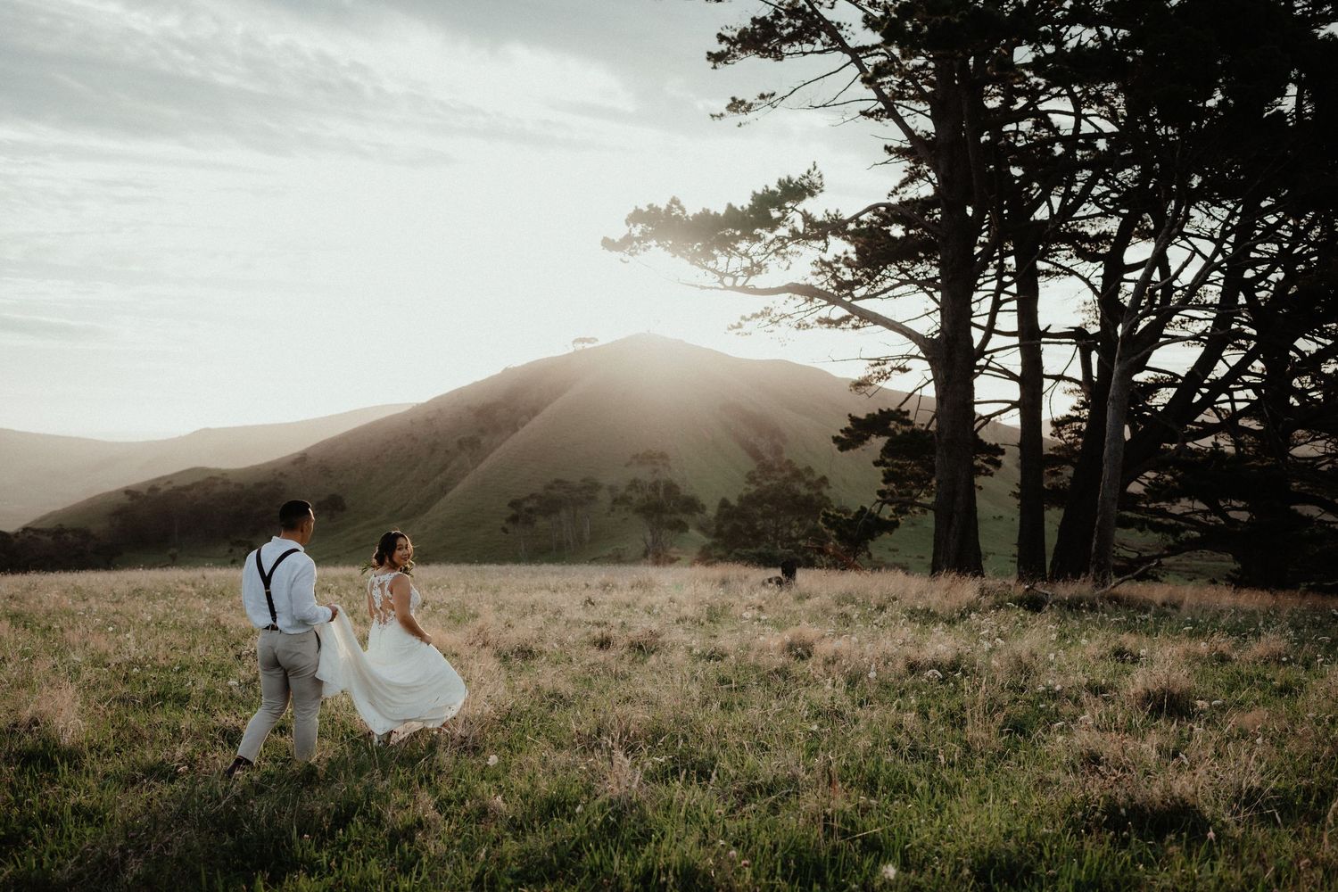 A bride and groom walk hand in hand through a grassy field at sunset with misty hills and cypress trees behind them.