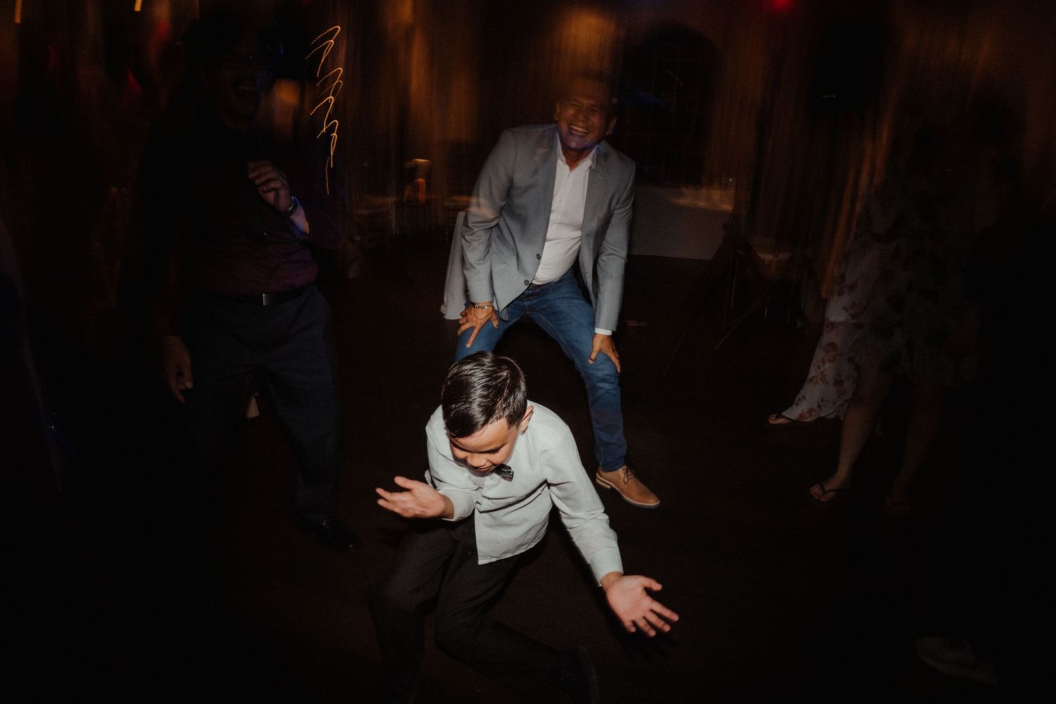 Guests dance energetically in a dark wedding reception, with a man in a light blazer and another dancing low in the foreground.
