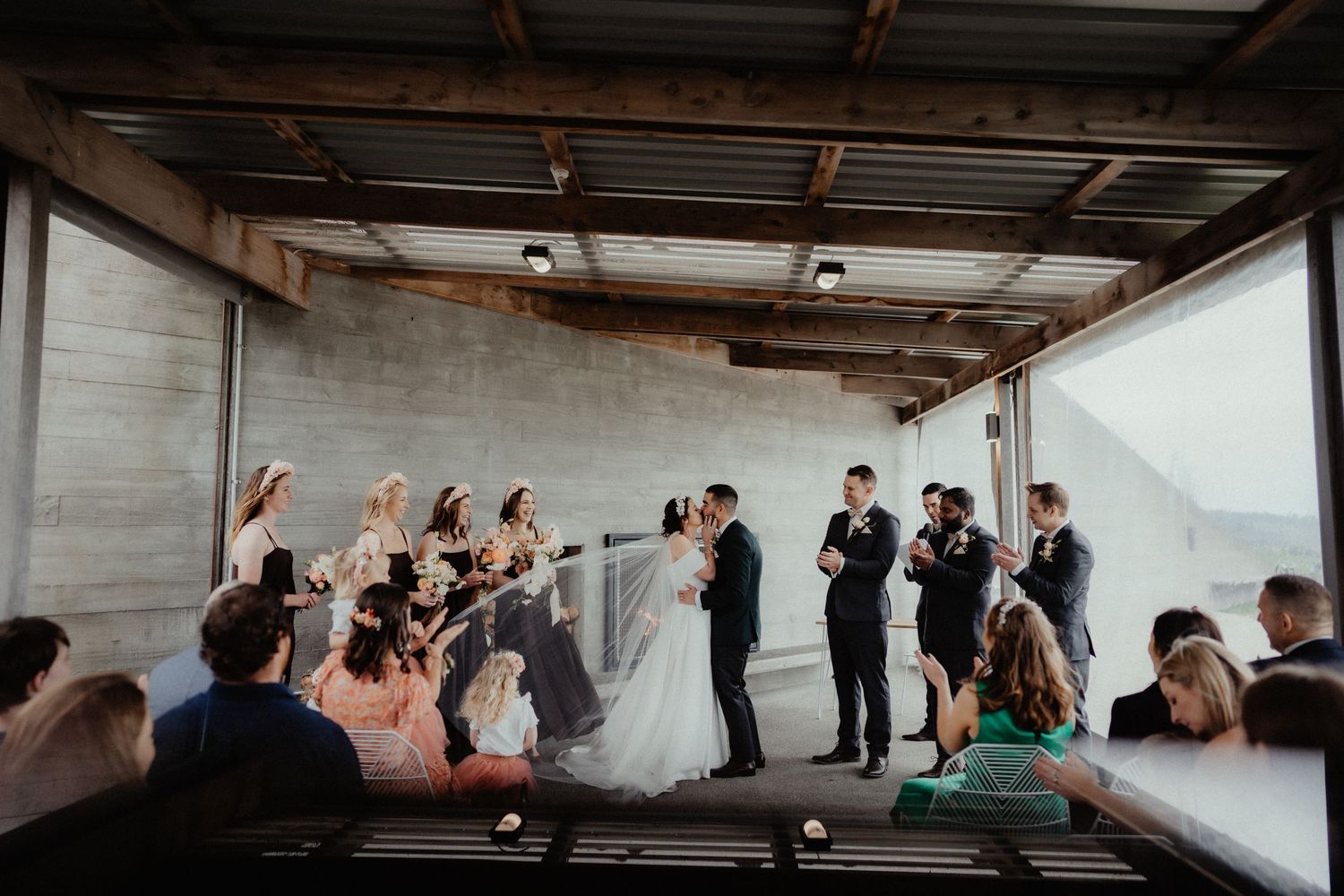 Couple kisses at the altar as bridesmaids, groomsmen and seated guests look on during a bright indoor wedding ceremony.