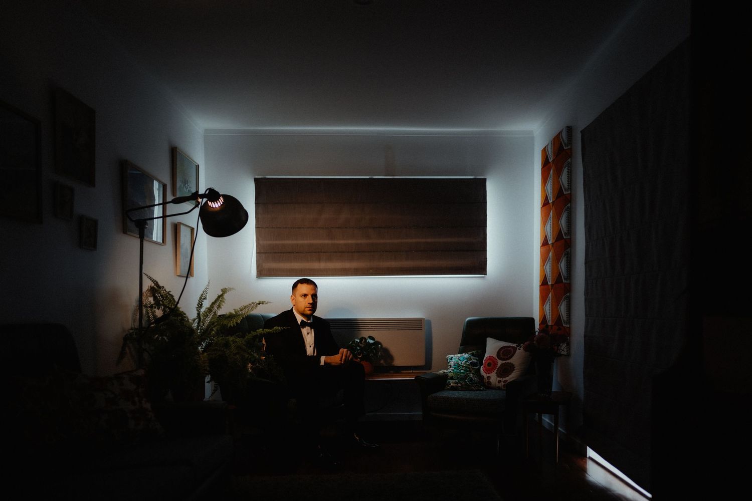 A man in a black suit and bow tie sits in a dimly lit living room, softly illuminated by window light and a floor lamp.