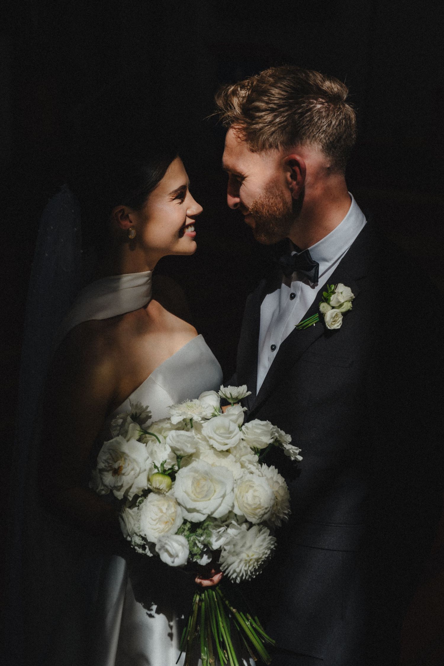 A bride and groom smile at each other in dramatic light, holding a bouquet of white roses.