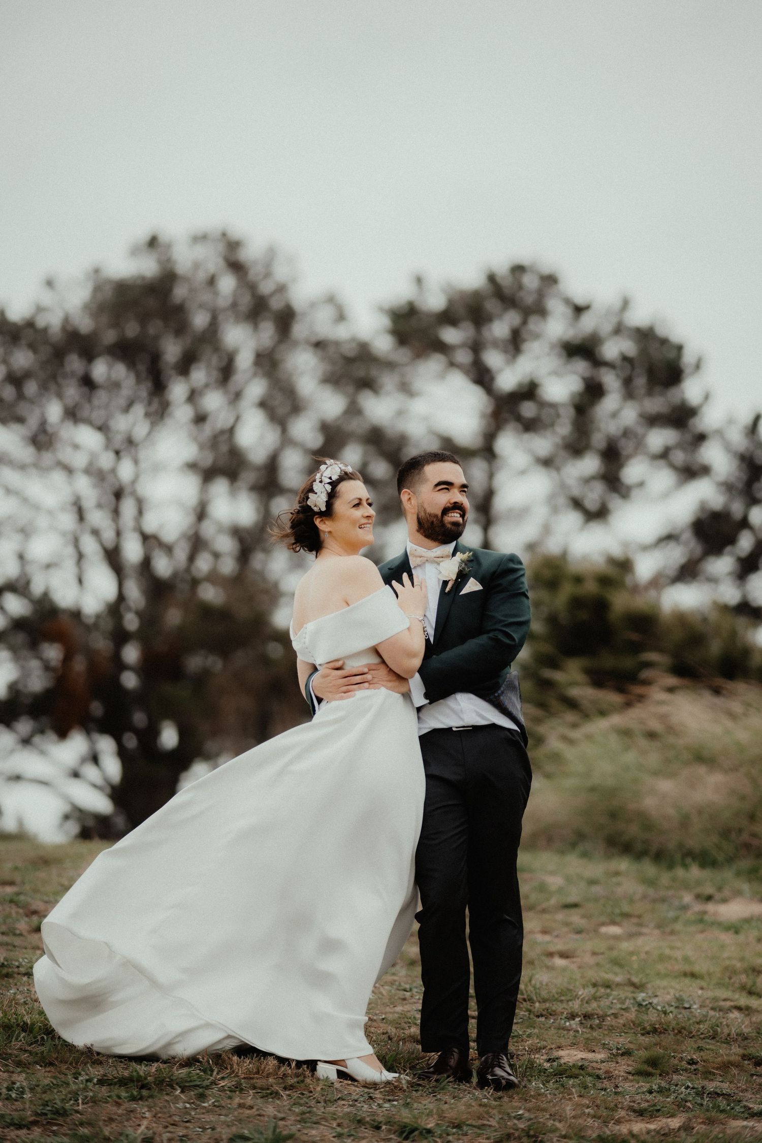 Bride in an off-shoulder white gown and groom in a dark suit embrace on a grassy hilltop on a windy overcast day.