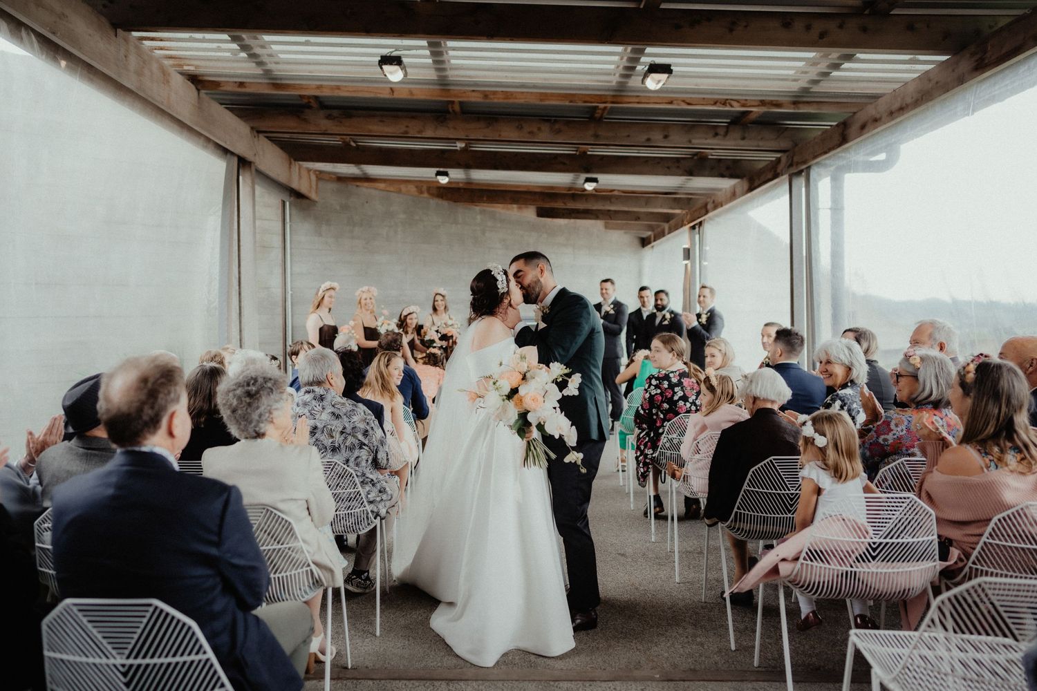 Bride and groom share a kiss at the end of the aisle as seated wedding guests watch during the ceremony.
