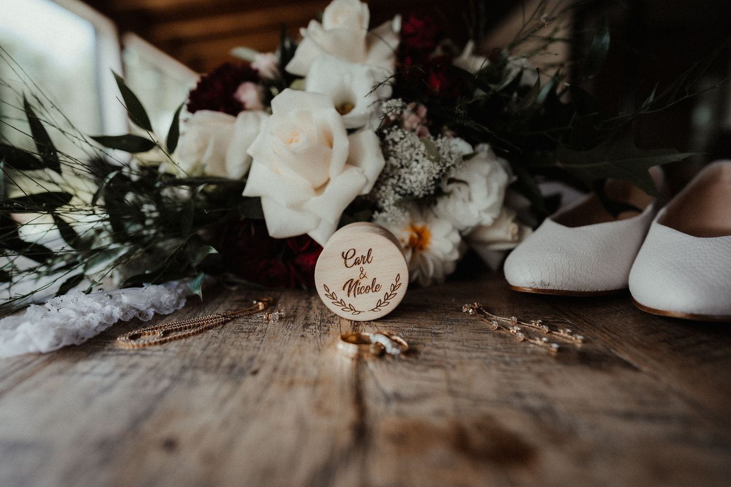 Wooden ring slice engraved with the couple's names rests on a rustic table beside white bridal shoes, wedding rings, and a floral bouquet.