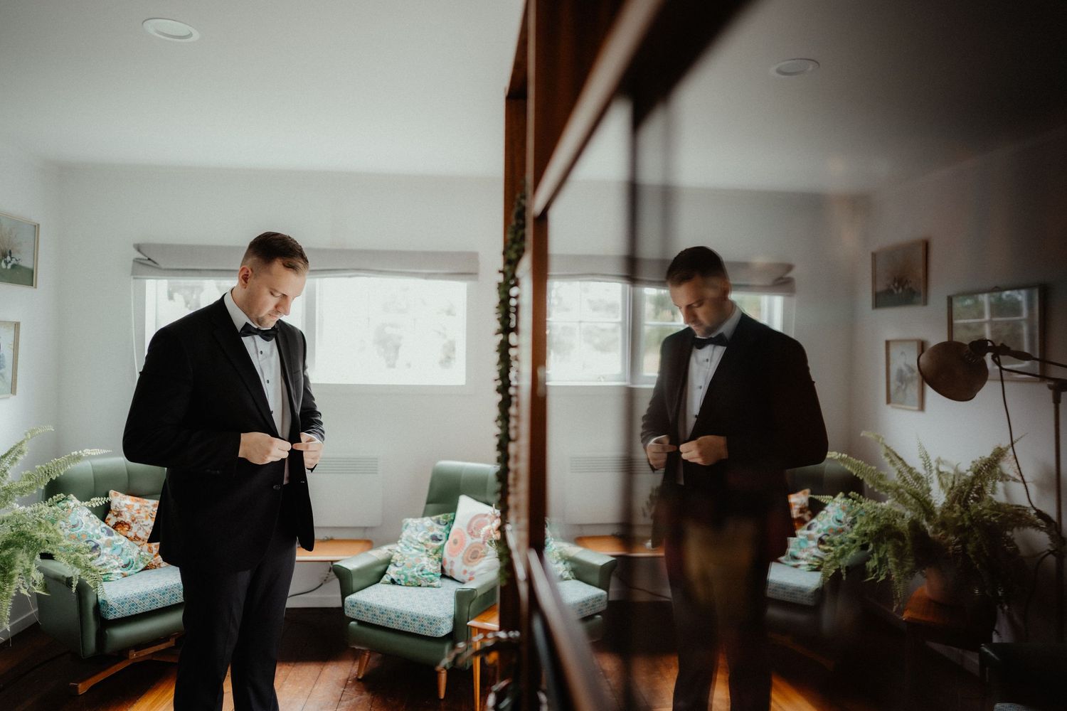 Groom in a black tuxedo and bow tie adjusts his jacket in a bright room with green armchairs, his reflection visible in a mirror.