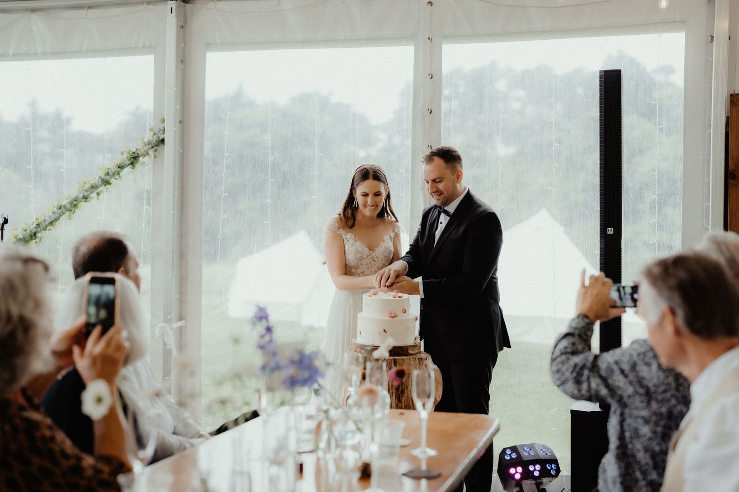 Bride and groom cut their tiered wedding cake together while guests photograph them with phones inside a marquee.