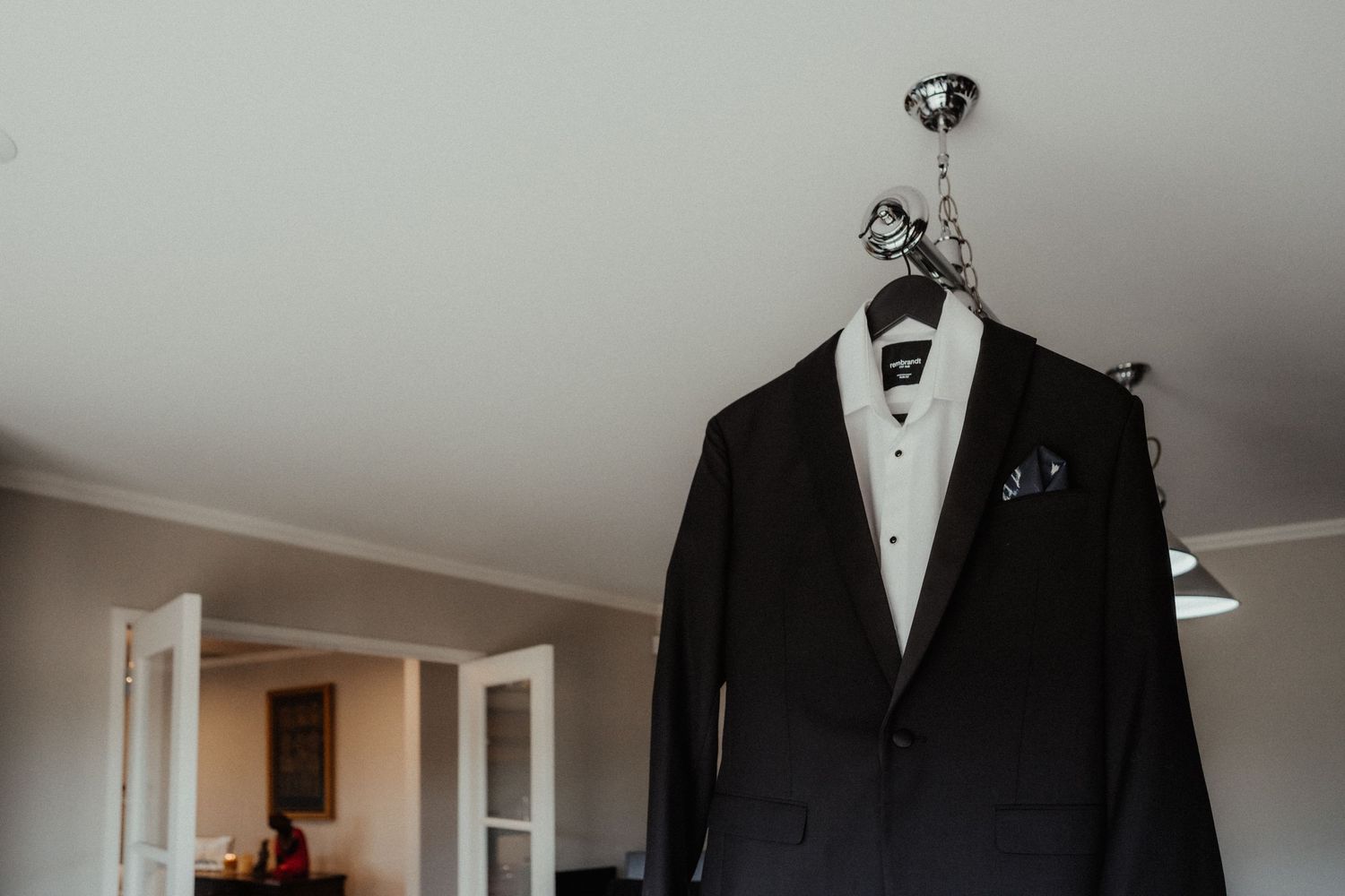 Black tuxedo jacket with a white shirt and pocket square hangs from a light fixture, ready for the groom on his wedding day.