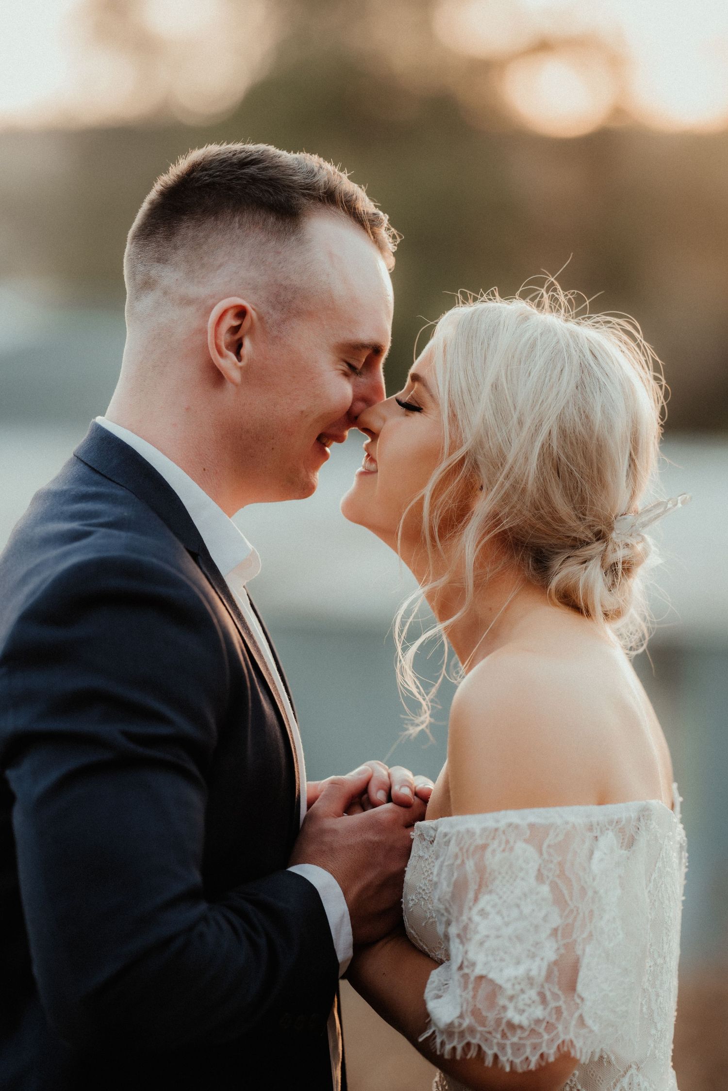 Smiling bride and groom touch noses and hold hands in warm golden light, the bride wearing an off-shoulder lace wedding dress.