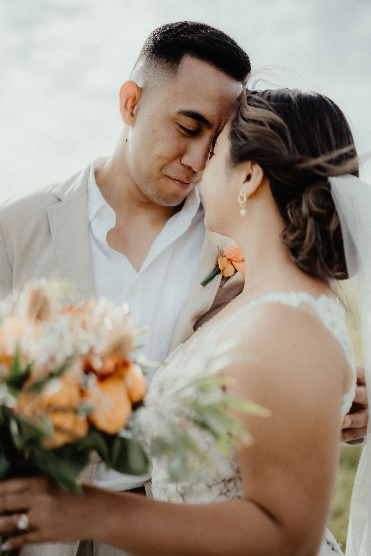 Groom in a beige linen jacket touches foreheads with his bride as she holds an orange and peach bridal bouquet outdoors.