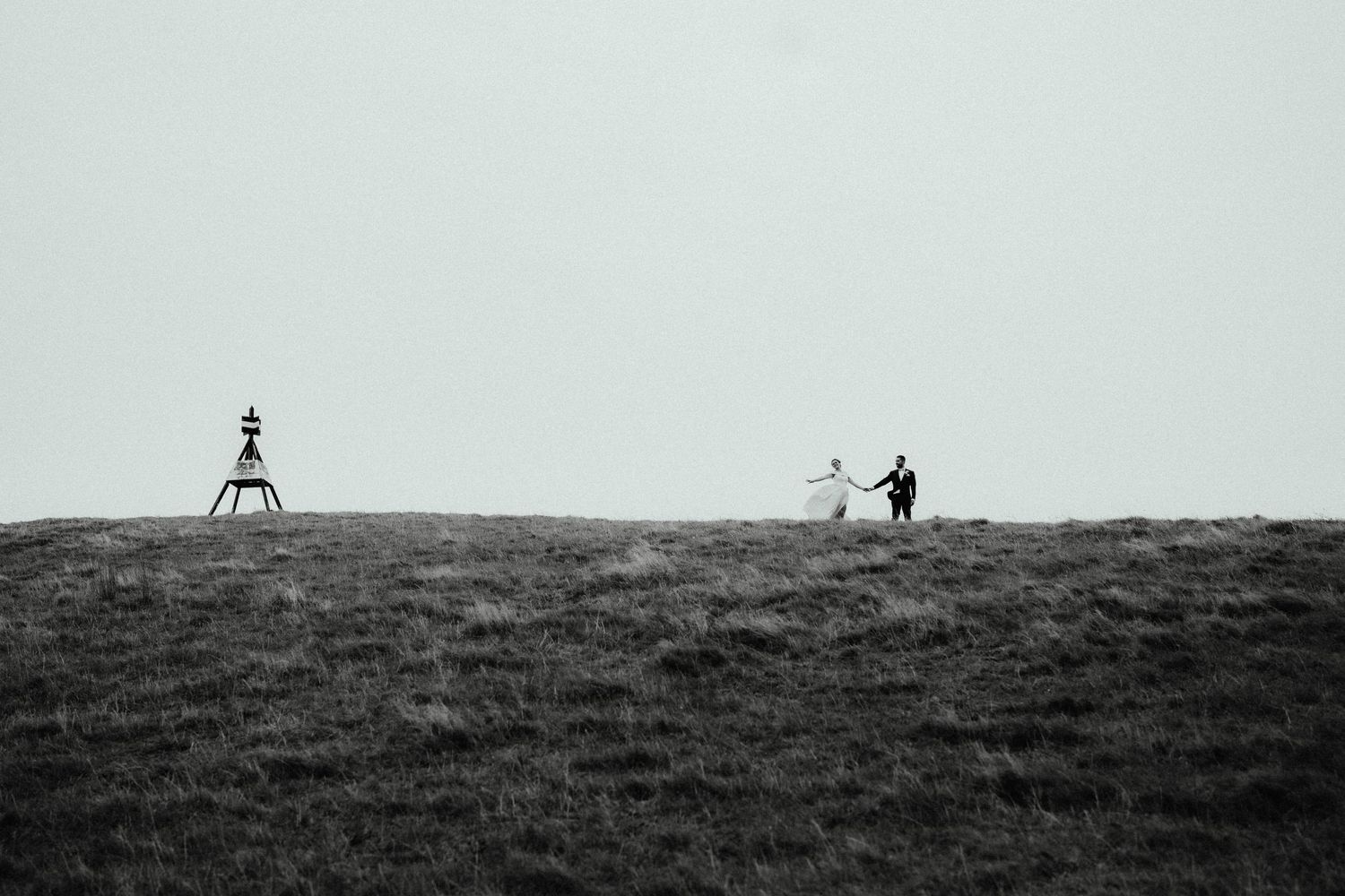 Bride and groom walk hand in hand across a grassy hilltop under an overcast sky in a black and white photo.