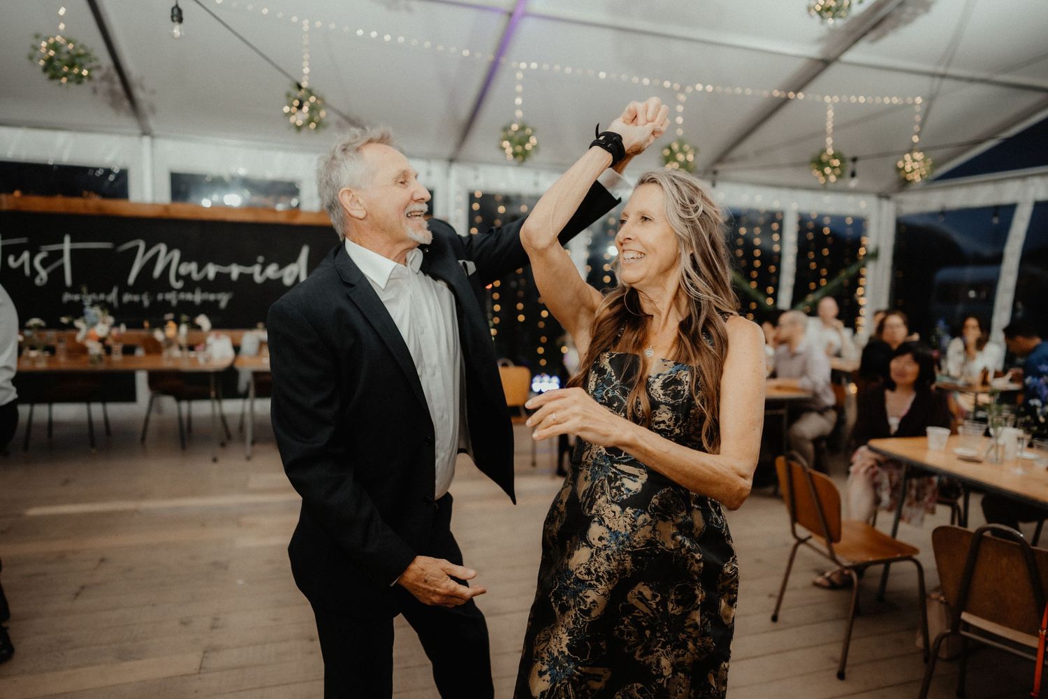 Smiling guest twirls a woman in a floral dress on the dance floor at a wedding reception with string lights.