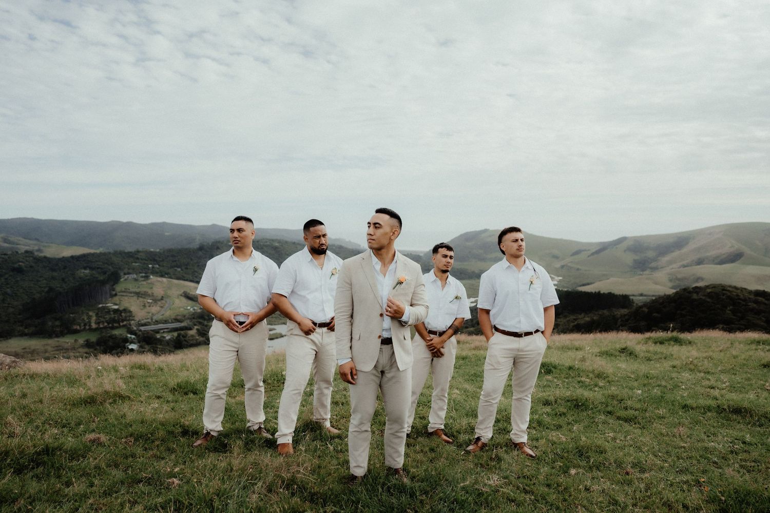 Groomsmen in beige suits stand on a grassy hillside with rolling green hills behind them at a countryside wedding.