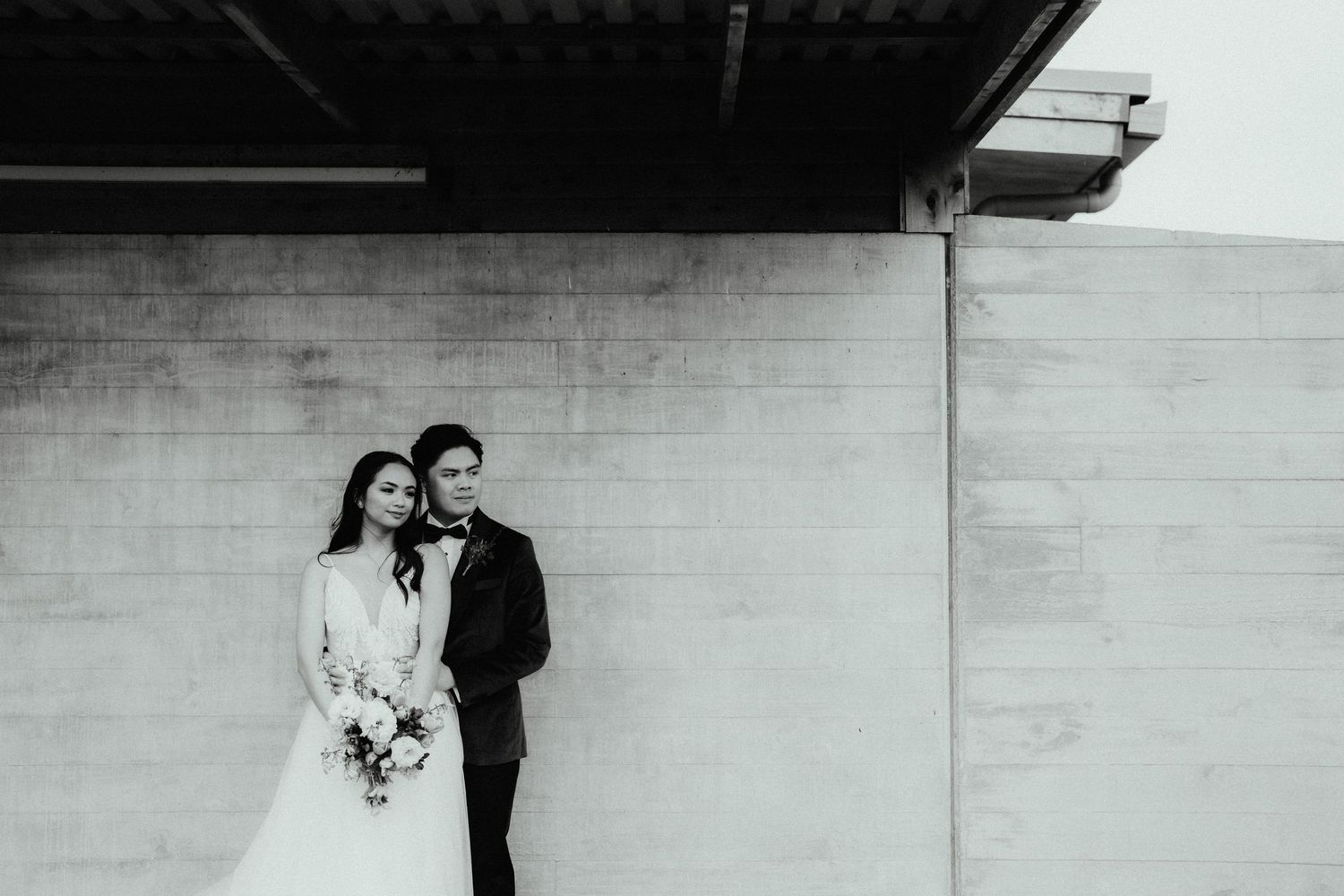 Black and white photo of a bride holding a bouquet leaning against her groom in front of a concrete wall.