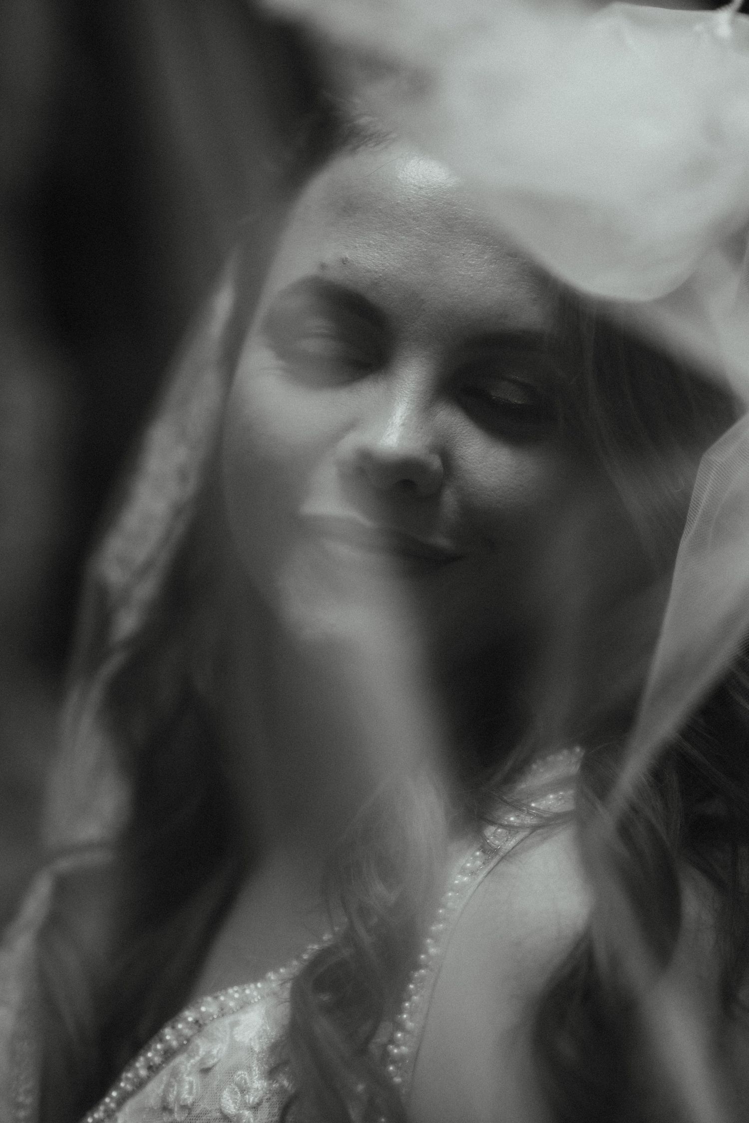 Soft black and white portrait of a smiling bride with closed eyes seen through a sheer veil.