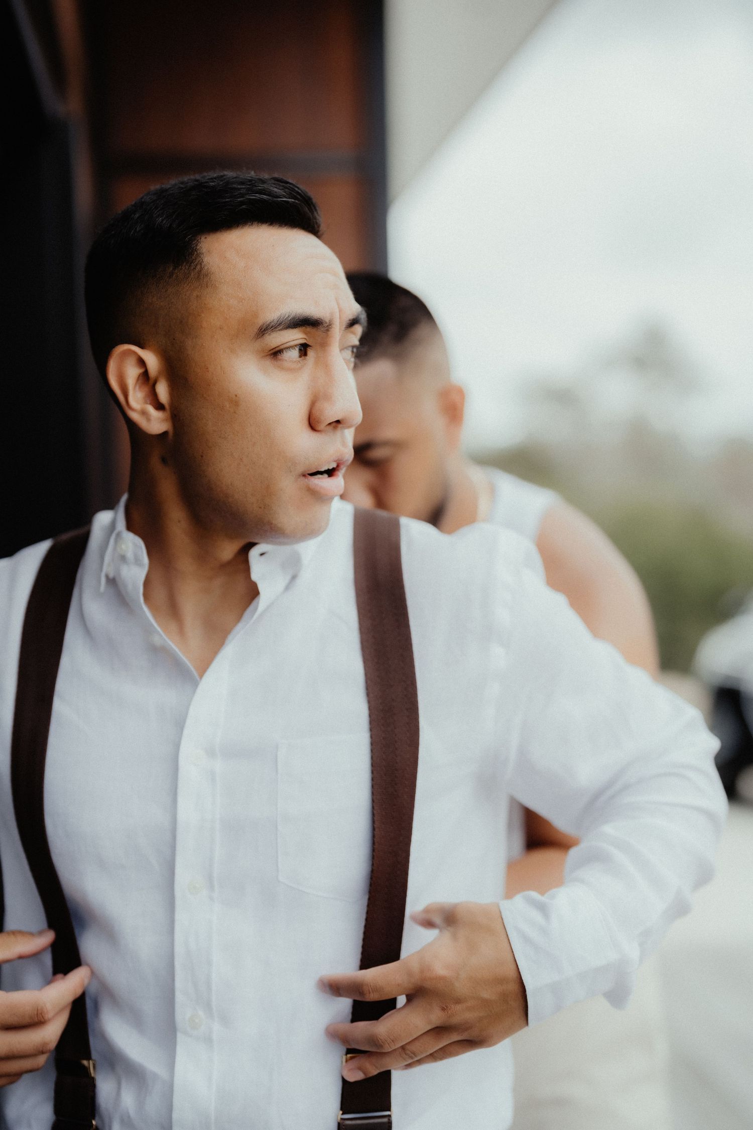 Groom in a white linen shirt and brown suspenders looks to the side while getting ready for his wedding with a groomsman behind him.