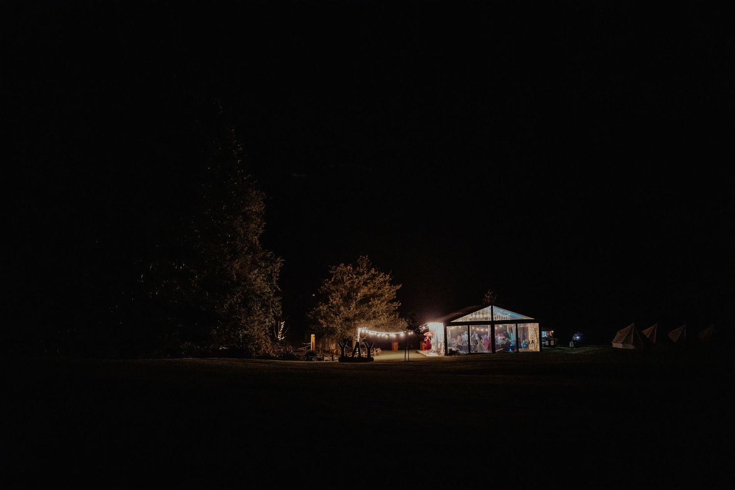A lit-up marquee glows in the dark countryside at night, surrounded by silhouetted trees, ahead of a wedding celebration.