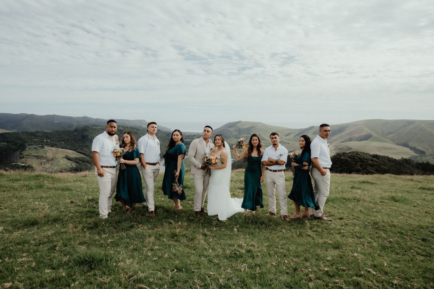 Wedding party in white shirts and teal dresses poses with the bride and groom on a grassy hilltop overlooking rolling hills.