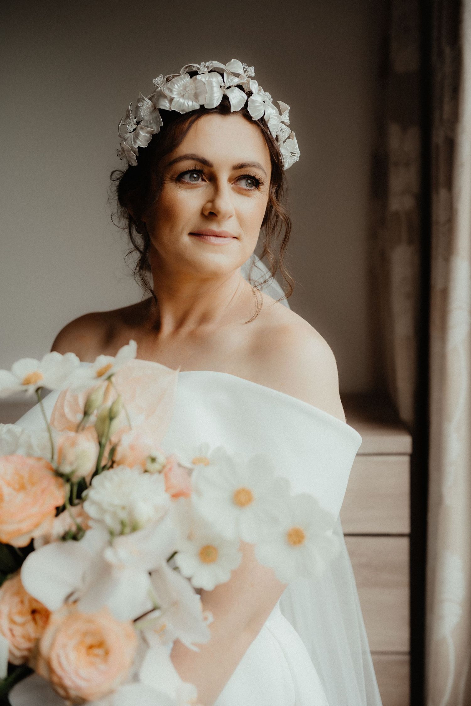 Bride wearing a floral headpiece and off-the-shoulder gown holds a bouquet of peach roses and white blooms beside a window.