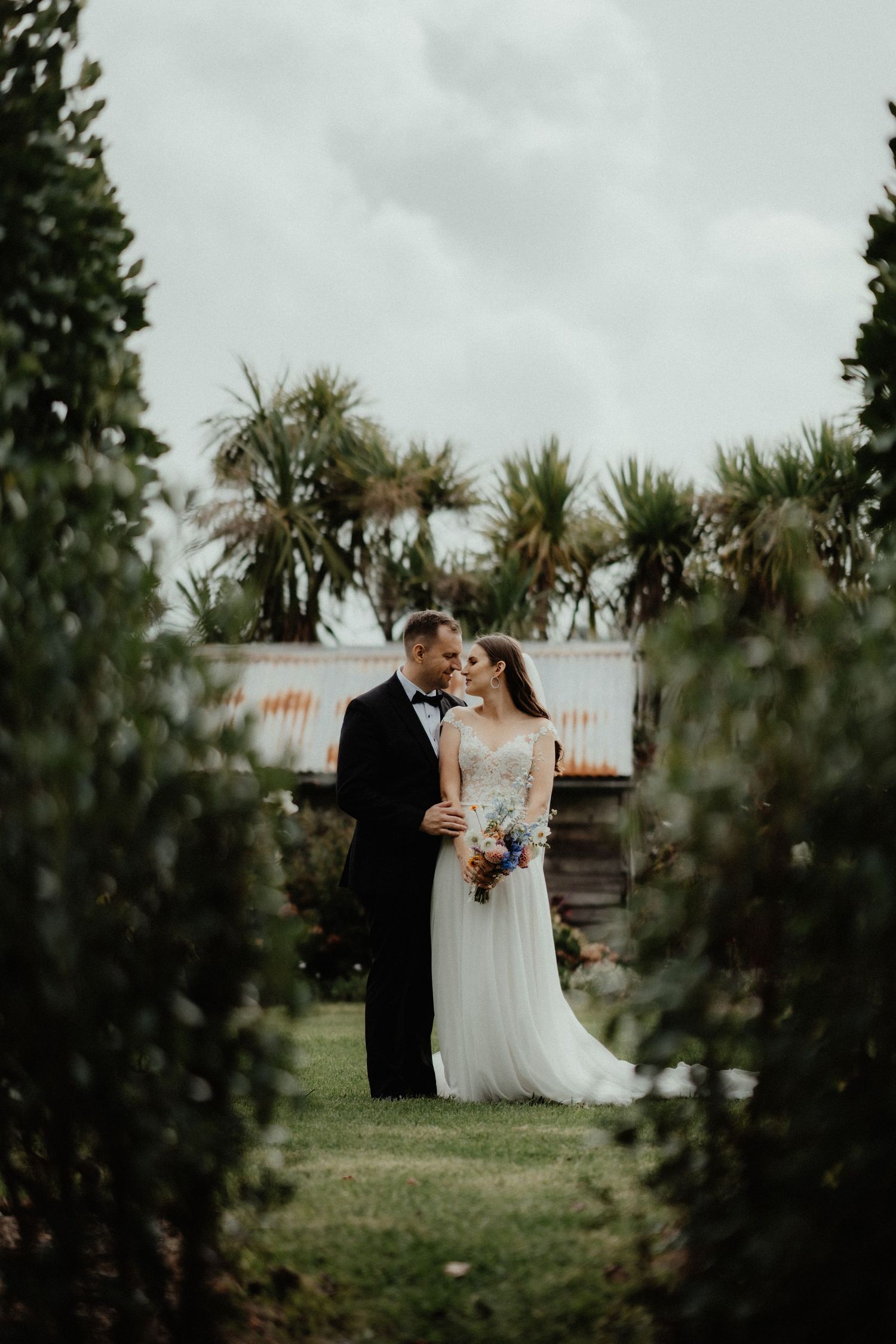 Bride and groom embrace on a green lawn between hedges, with palm trees and a rustic barn behind them.