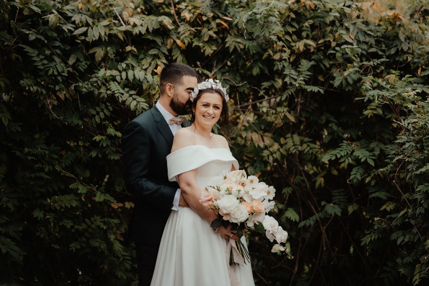 Groom kisses the cheek of his smiling bride, who wears a floral crown and holds a white bouquet in front of dense green foliage.