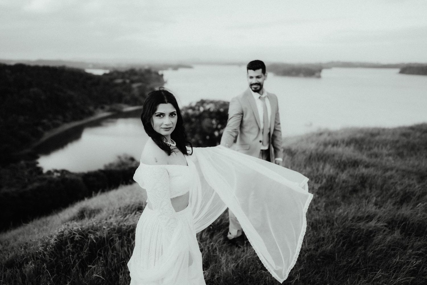 Black and white photo of a bride in a flowing sheer gown and a groom on a grassy hilltop overlooking a calm bay.