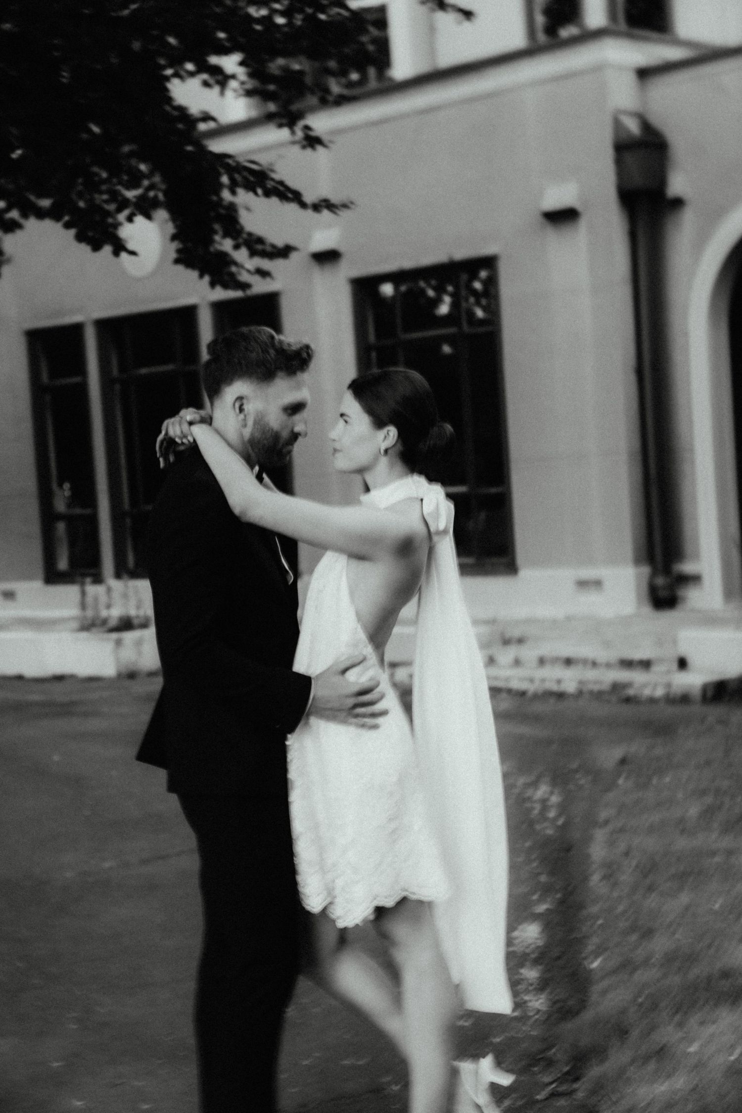 Black and white photo of a groom in a dark suit embracing a bride in a short lace wedding dress with a bow outside a building.