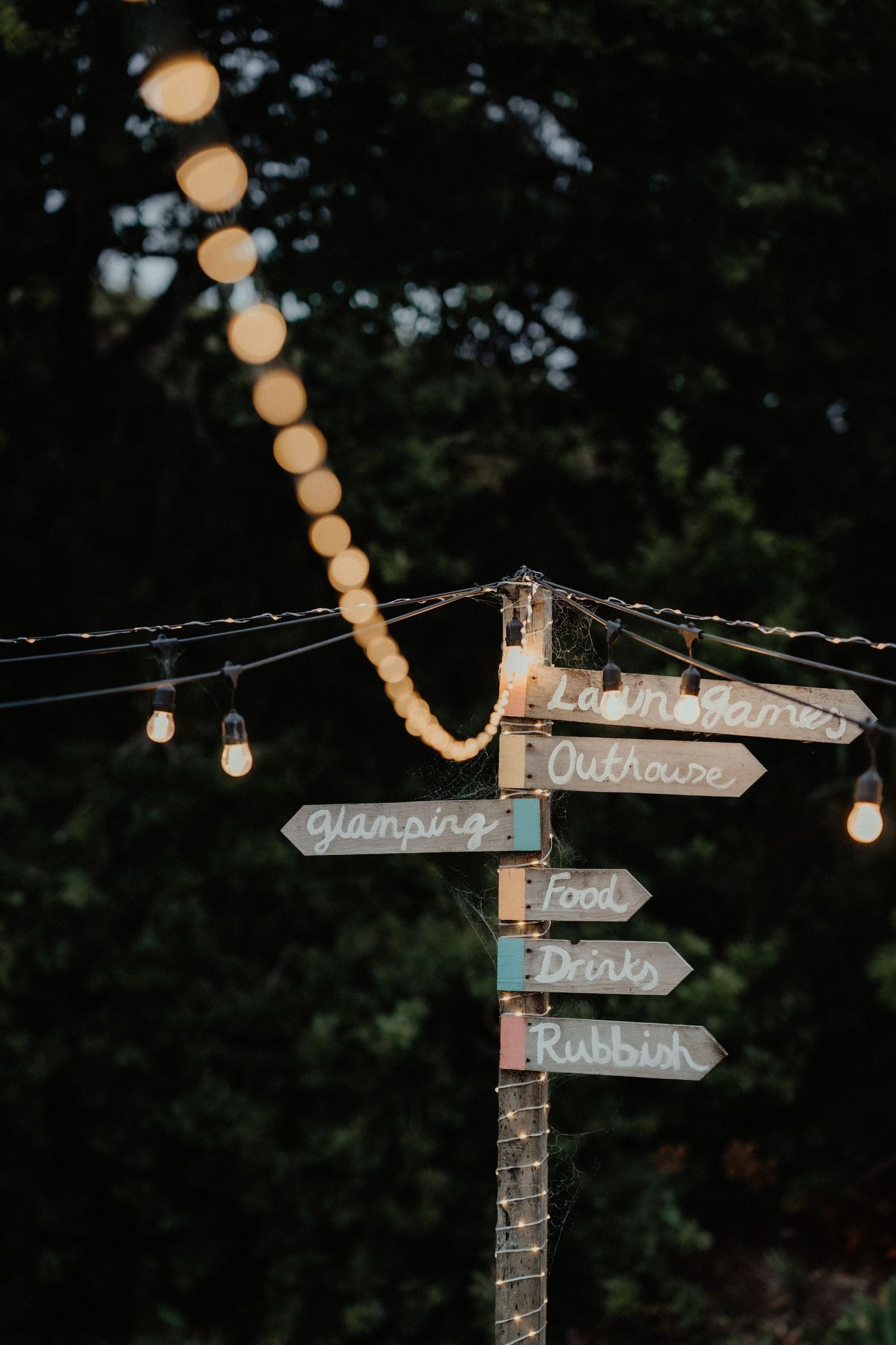 A wooden signpost strung with glowing festoon lights points to glamping, outhouse, food, drinks and rubbish at a evening wedding.
