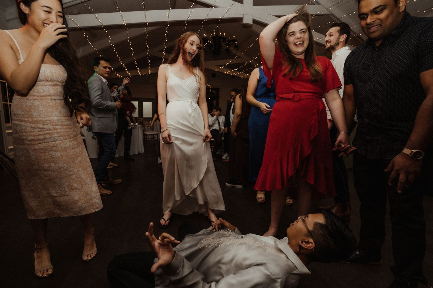 Wedding guests laugh and celebrate on the dance floor while a man lies on the ground in the foreground during the reception.
