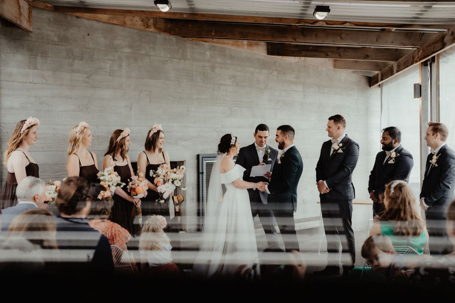 Bride and groom hold hands during an outdoor-style wedding ceremony as bridesmaids and groomsmen look on in an industrial venue.