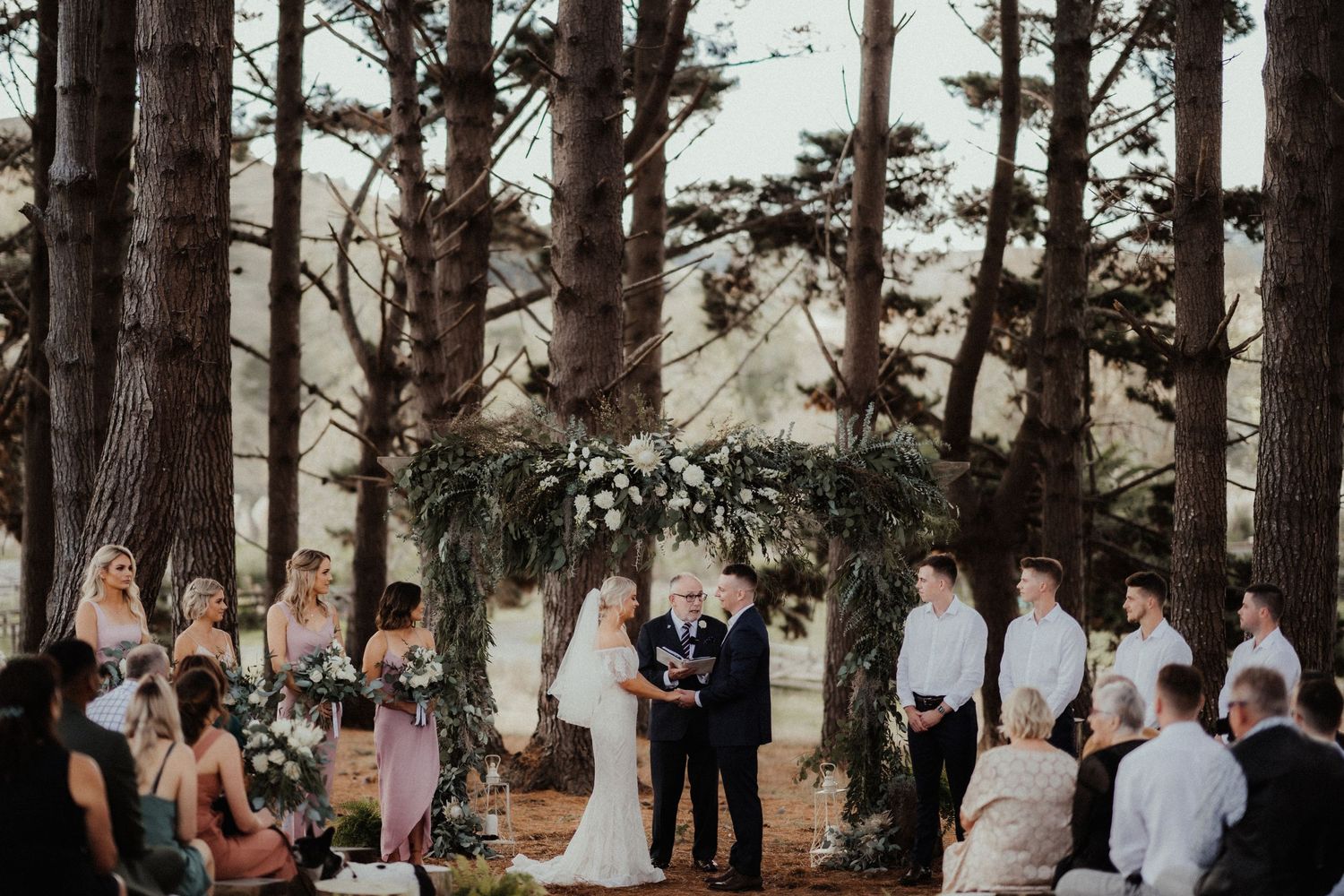 Couple exchange vows beneath a rustic floral arch in a pine forest as bridesmaids, groomsmen and guests look on.
