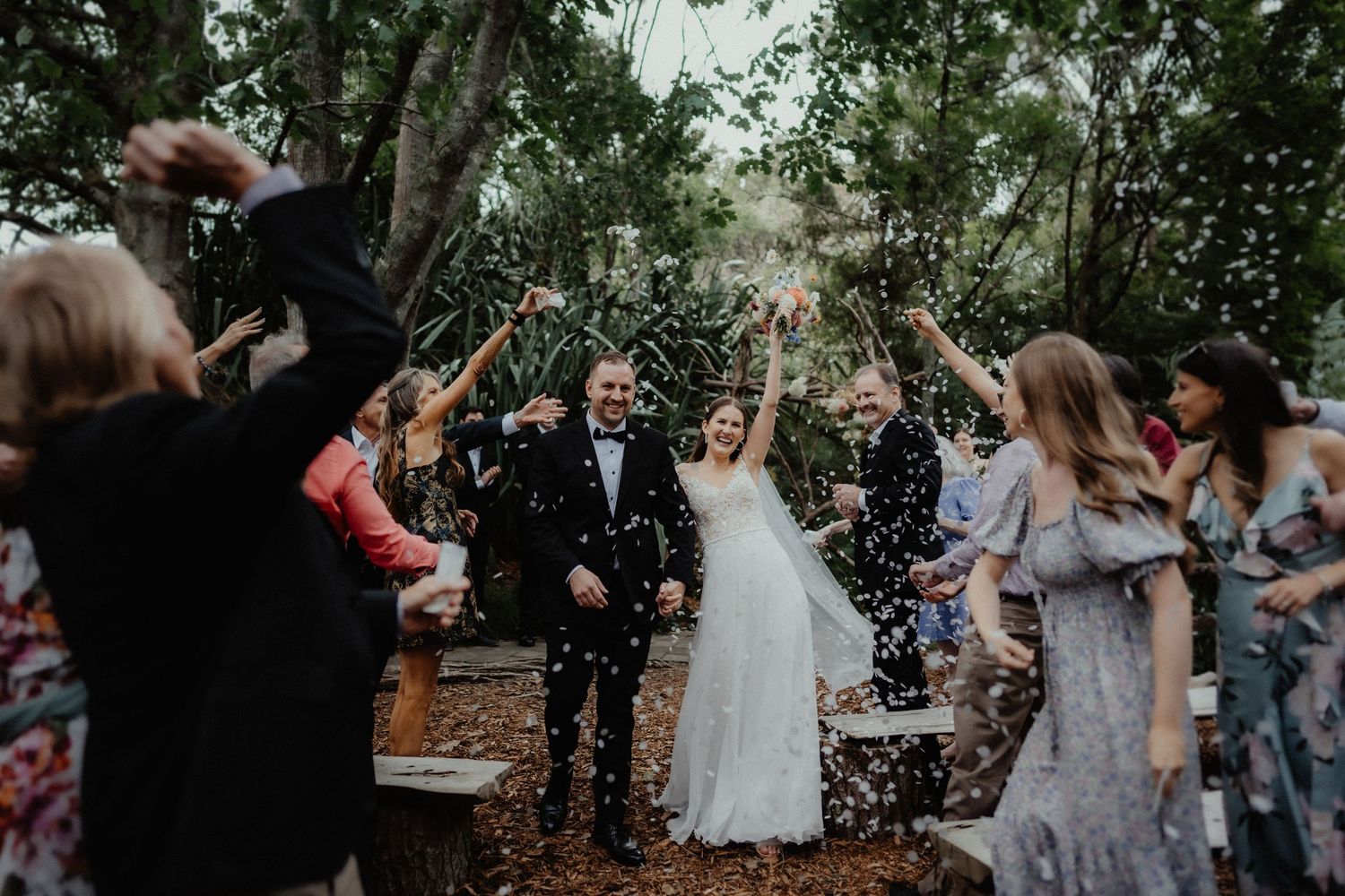 Joyful bride and groom raise their arms as guests throw confetti along a wooded aisle after the wedding ceremony.