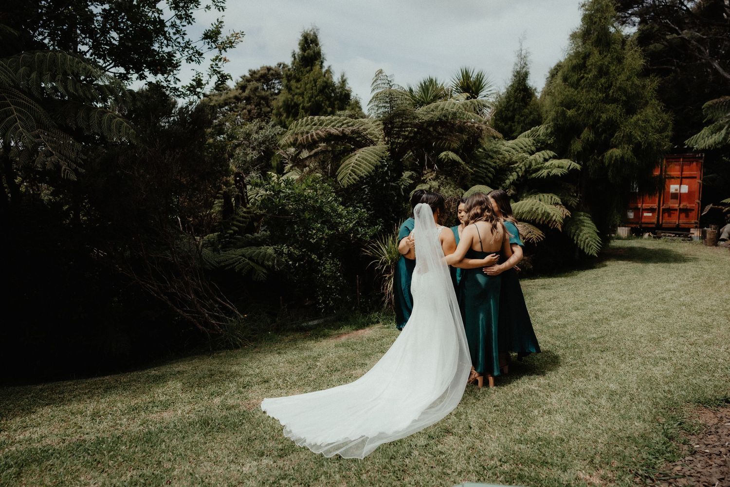 Bride in a long-train white gown is attended by bridesmaids in emerald green dresses beside lush ferns.
