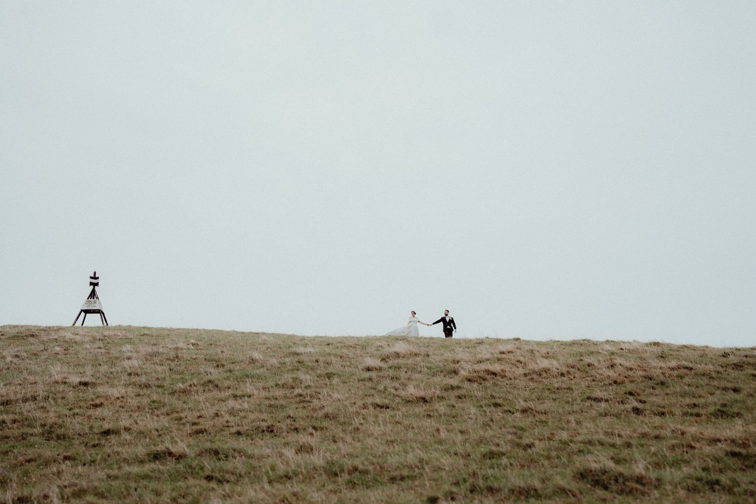Bride and groom walk together across a wide, misty grass hilltop beneath a pale grey sky, with a small tower nearby.