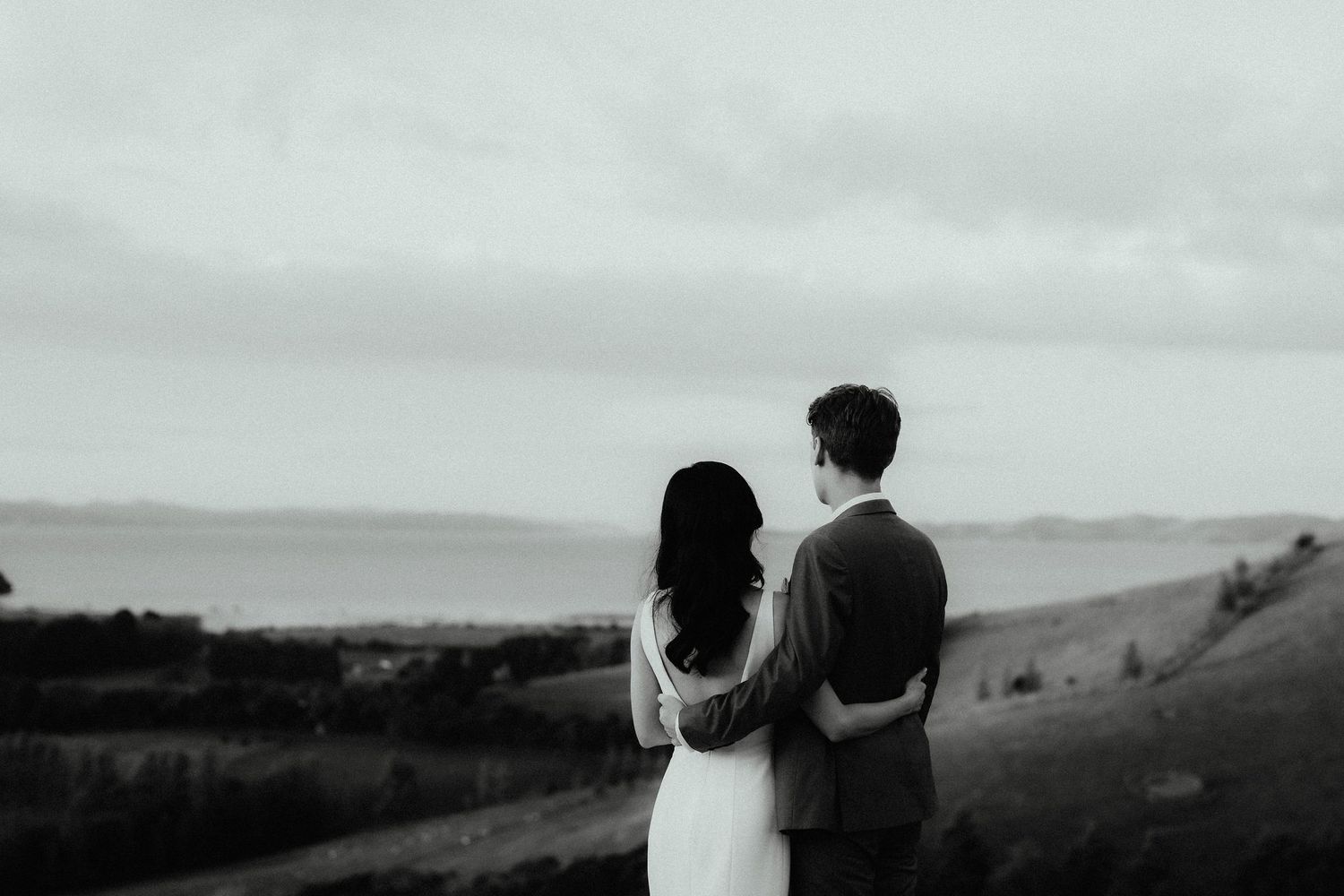 Black and white photo of a bride and groom embracing from behind as they gaze over rolling hills toward the sea.