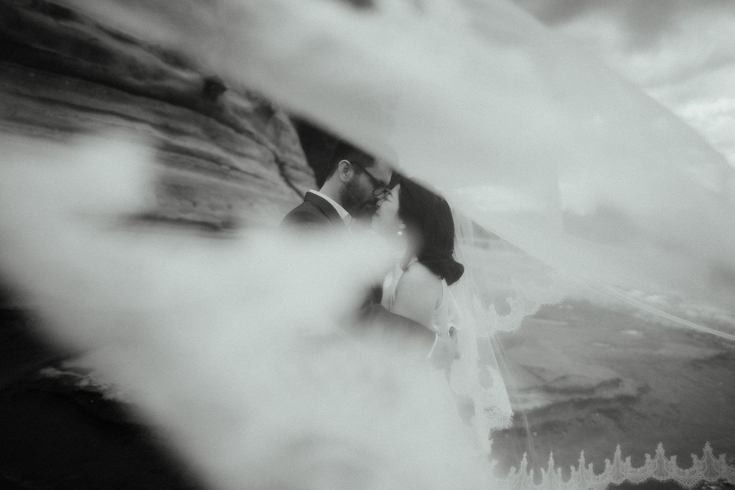 Dreamy black and white wedding photo of a couple kissing beneath a flowing veil with soft, misty movement.