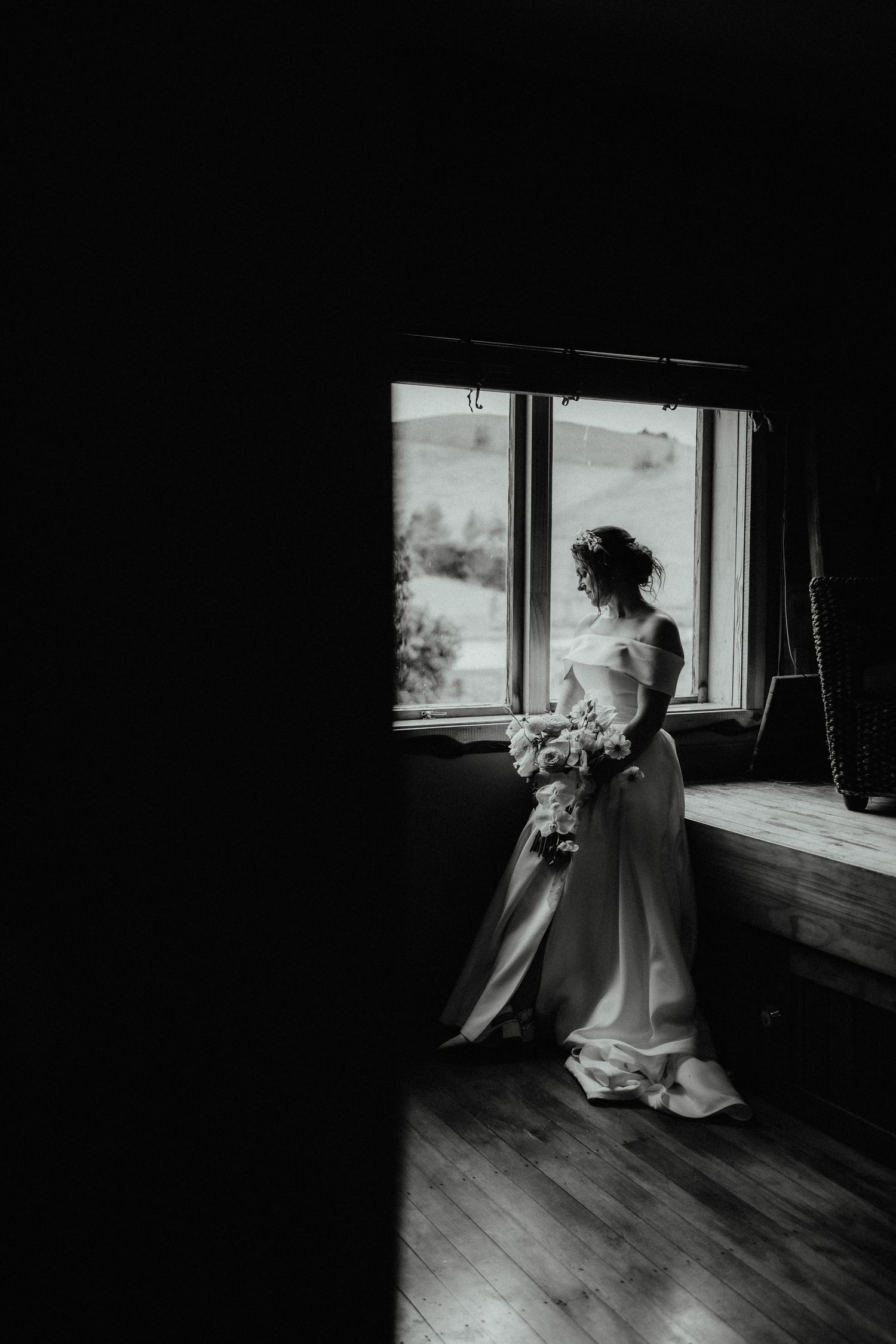 Black and white photo of a bride in an elegant satin gown holding a bouquet beside a window in a dim room.