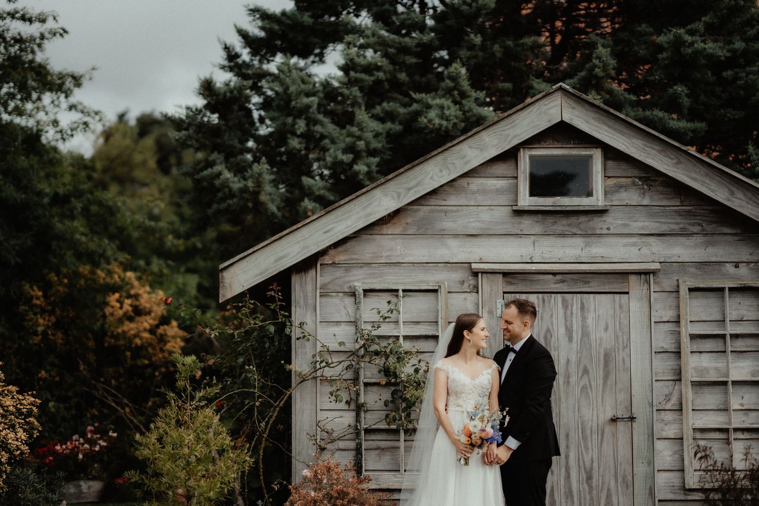 A bride and groom embrace in front of a weathered wooden shed surrounded by autumn trees and garden greenery on their wedding day.