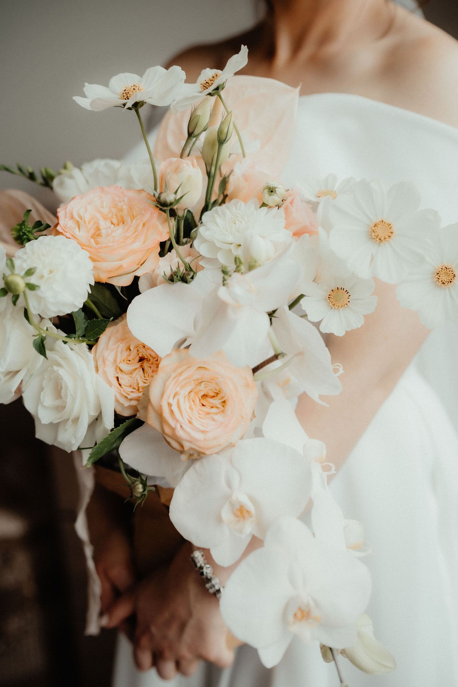 Bride in a white gown holds a romantic bouquet of peach roses, white orchids and daisies.