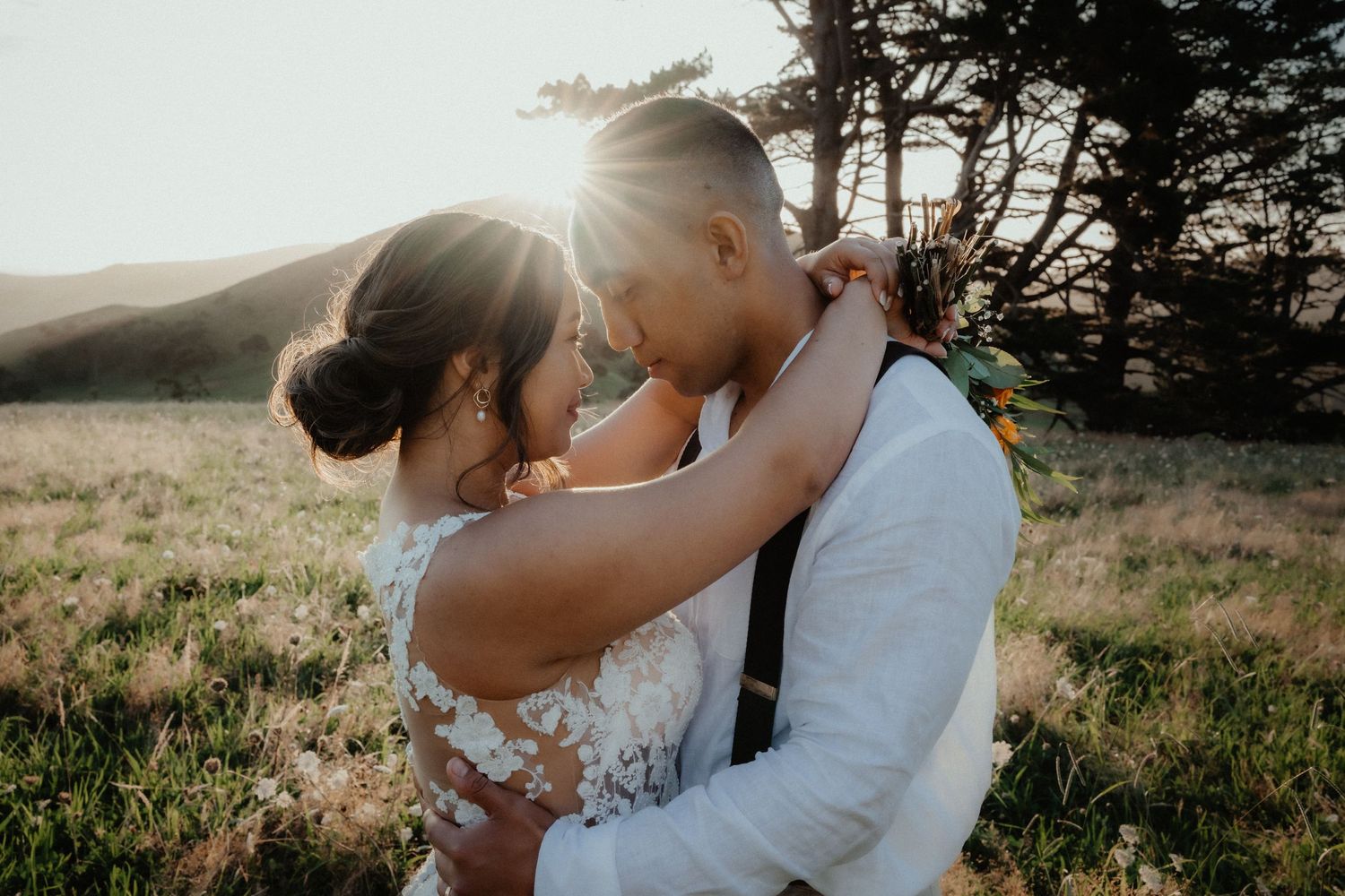 Bride and groom embrace in a grassy field as golden sunlight glows behind them at an outdoor wedding photoshoot.