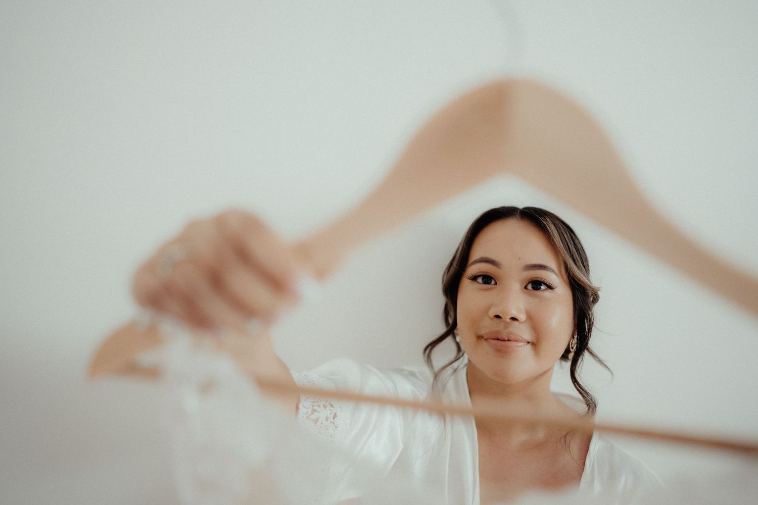 Smiling bride in a white robe holds a wooden hanger with her wedding dress while getting ready.