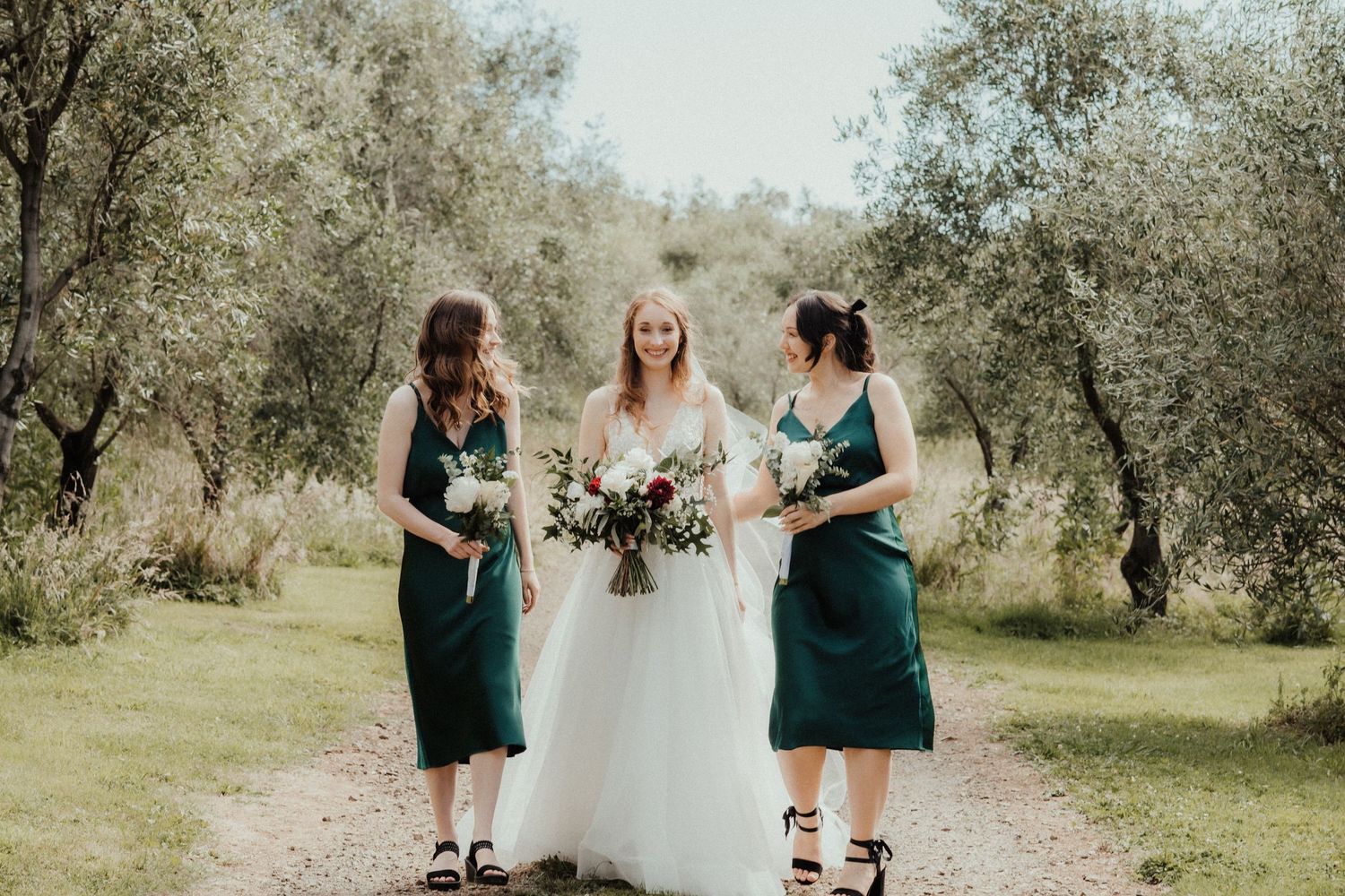A bride walks along an olive grove path with two bridesmaids in green dresses, all holding floral bouquets.