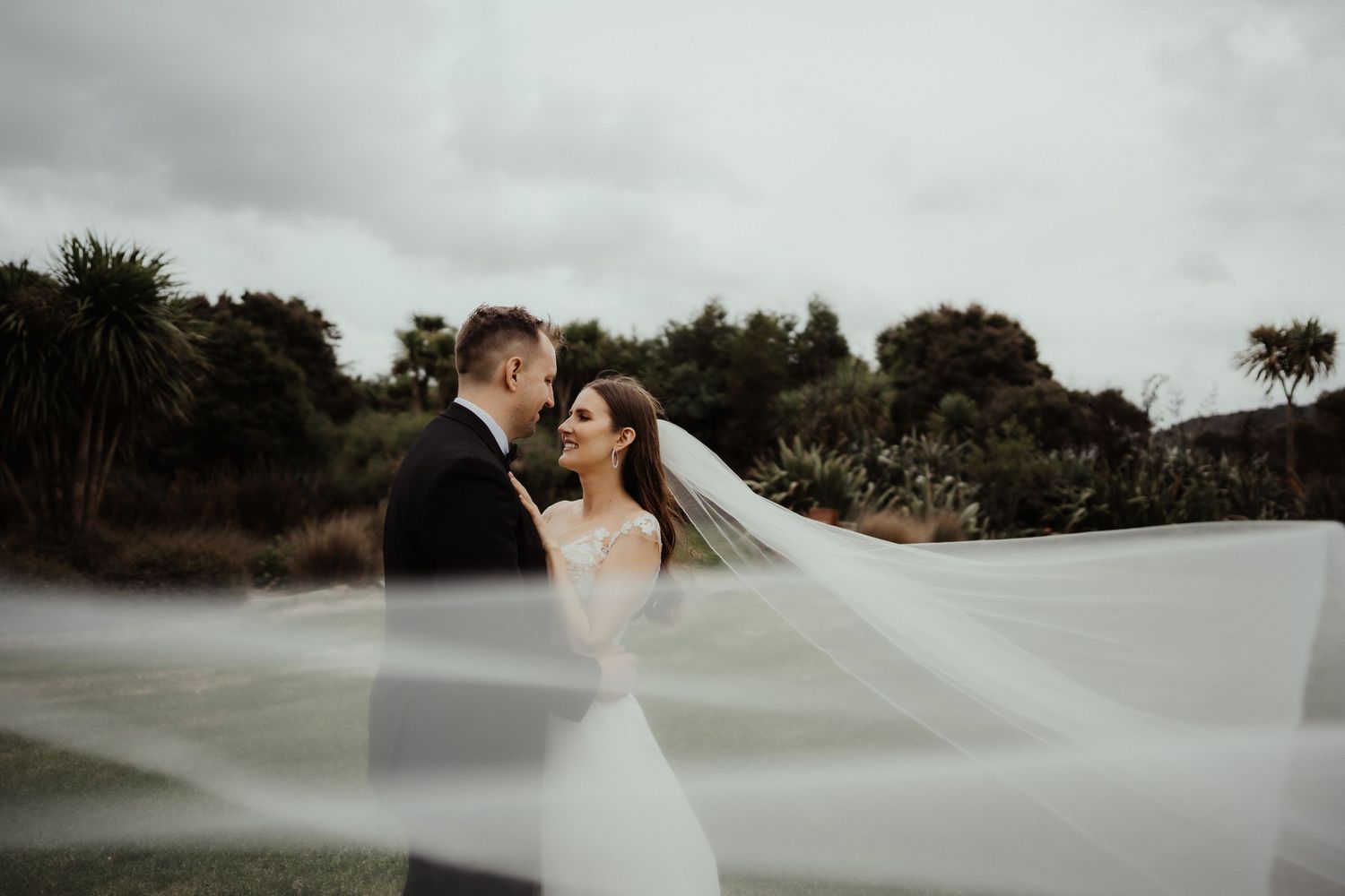 Bride and groom embrace outdoors as the bride's long sheer veil floats in the wind against a backdrop of trees.