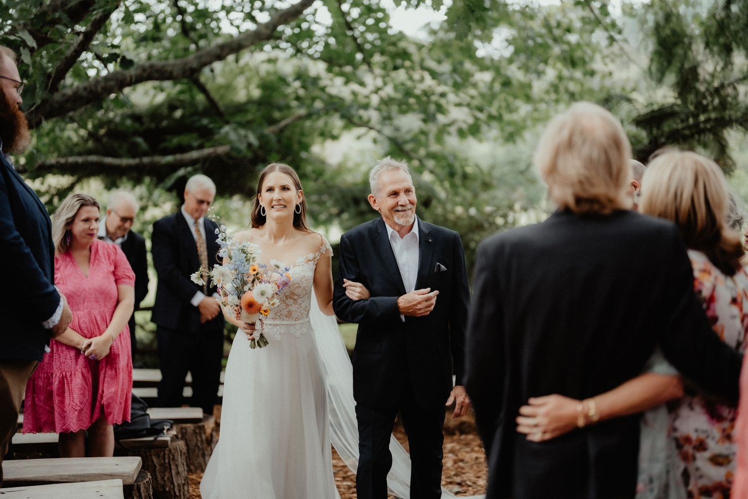 Bride in a white gown carrying a bouquet walks down the outdoor aisle on her father's arm as guests watch under the trees.