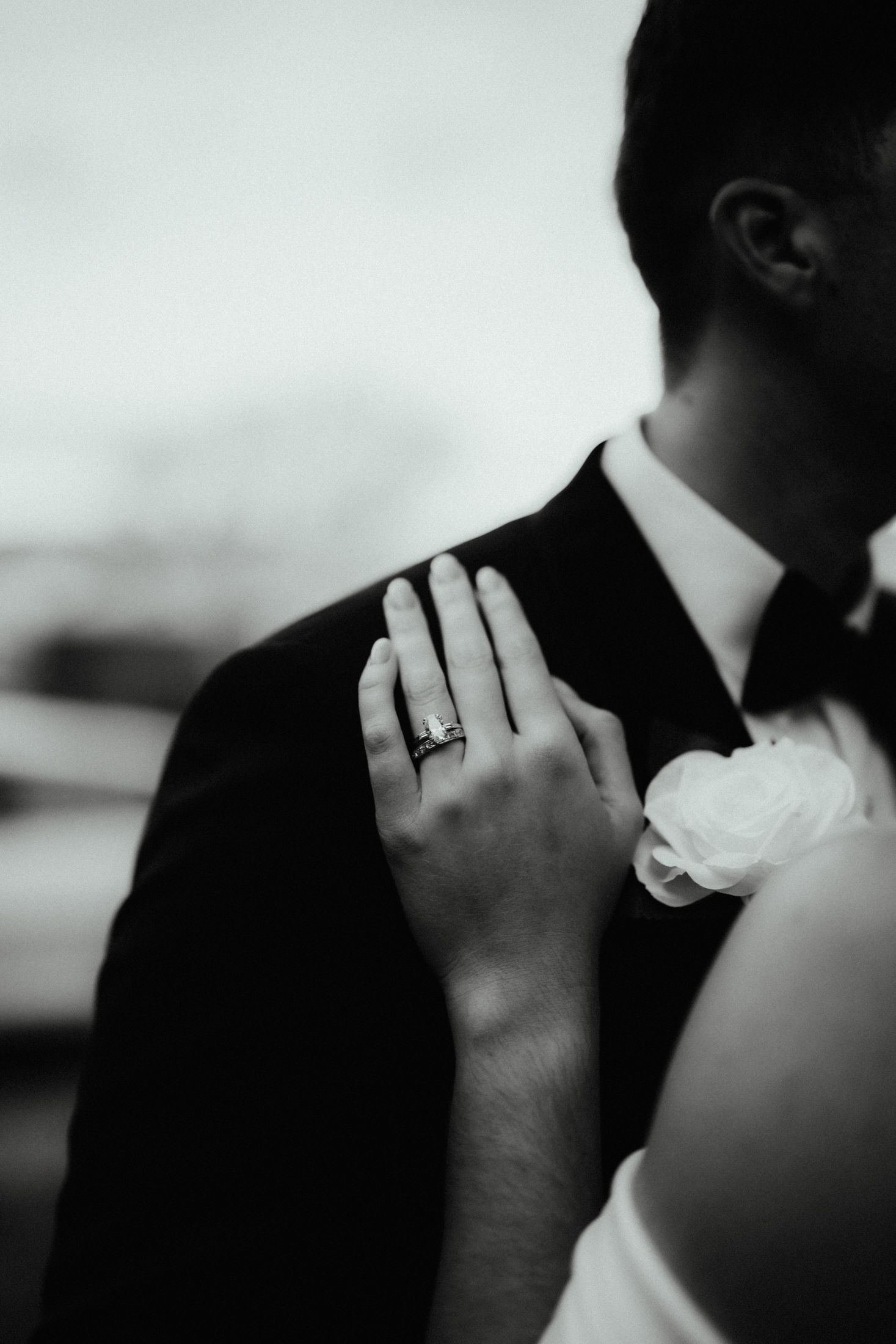 Black and white close-up of a bride's hand with a diamond ring resting on her groom's shoulder near a boutonniere.