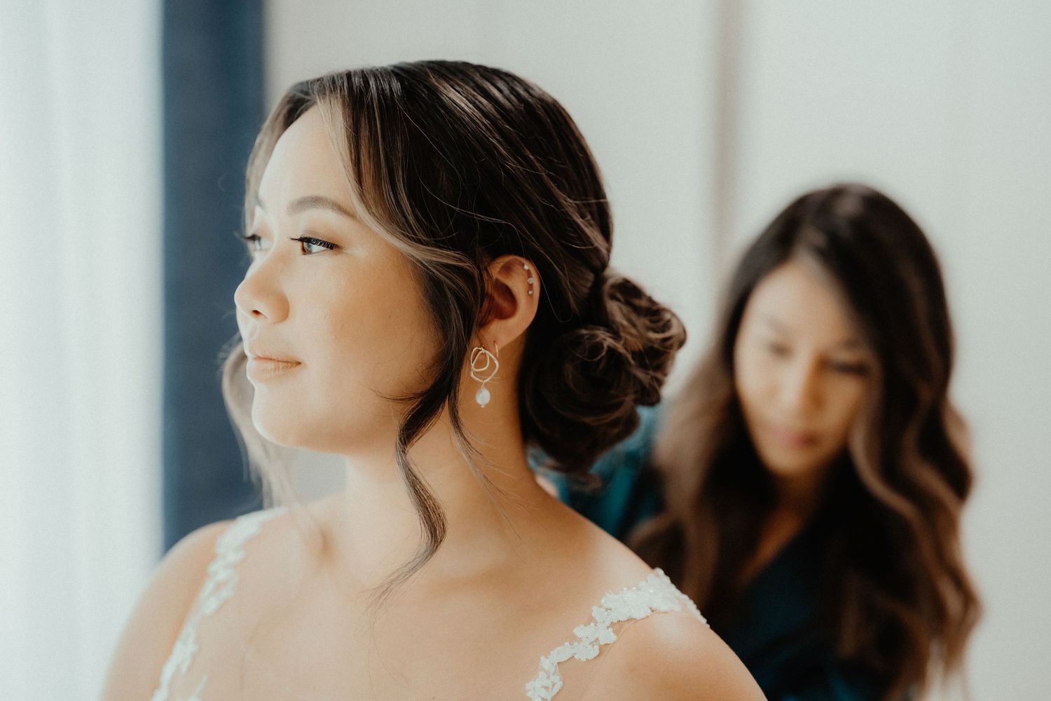 A bride with an elegant updo and drop earrings looks aside while a woman helps her get ready for the wedding.