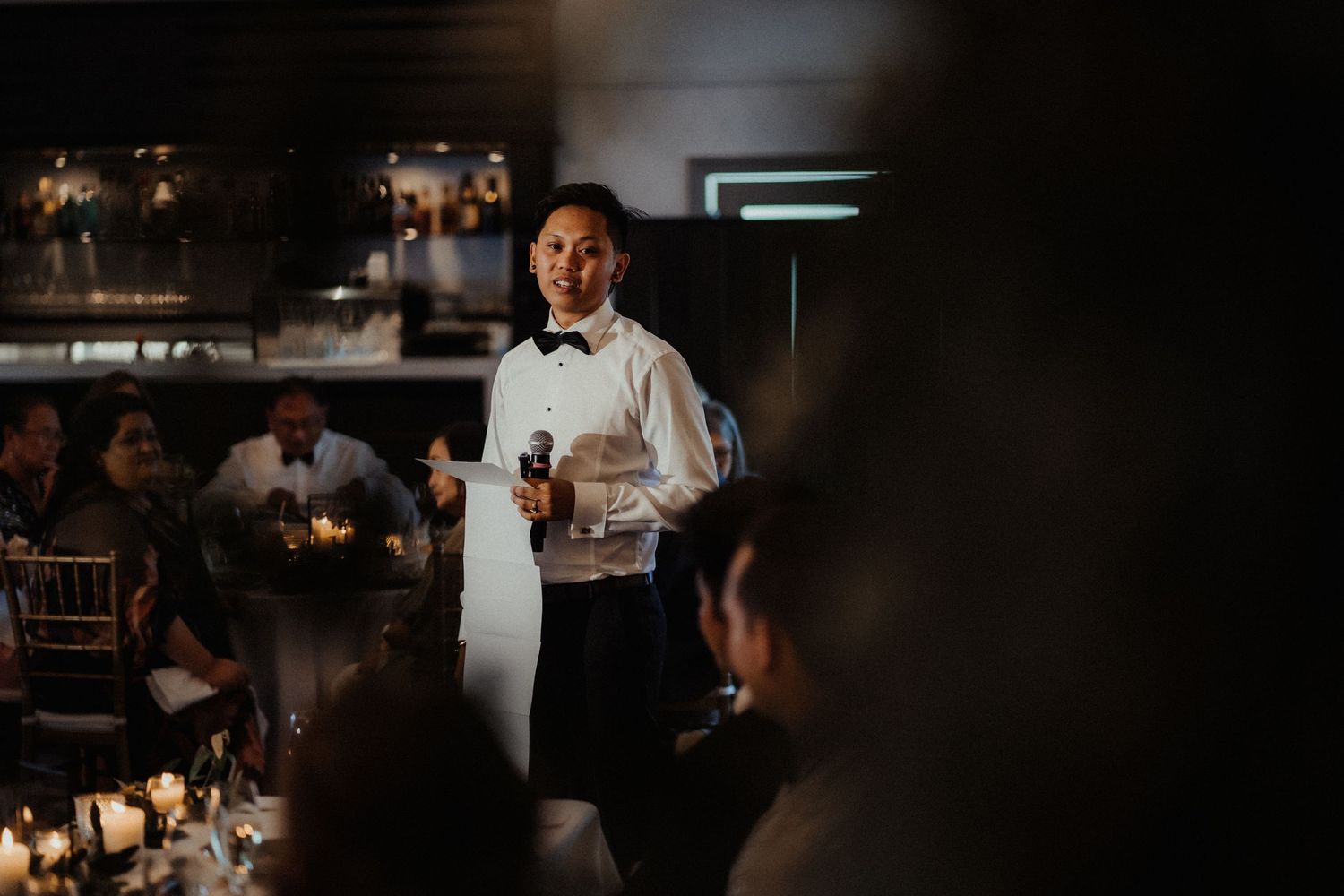 Man in a white shirt and bow tie speaks into a microphone at a candlelit wedding reception as guests listen at the tables.