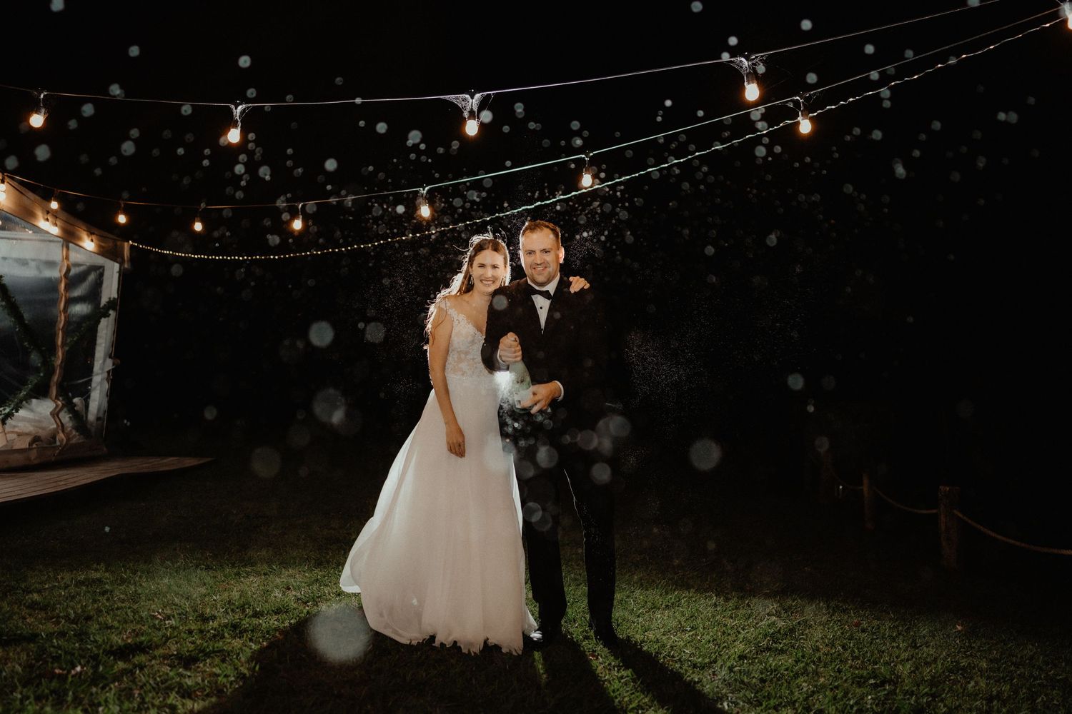 Bride and groom embrace on a lawn at night beneath glowing string lights during their wedding reception.
