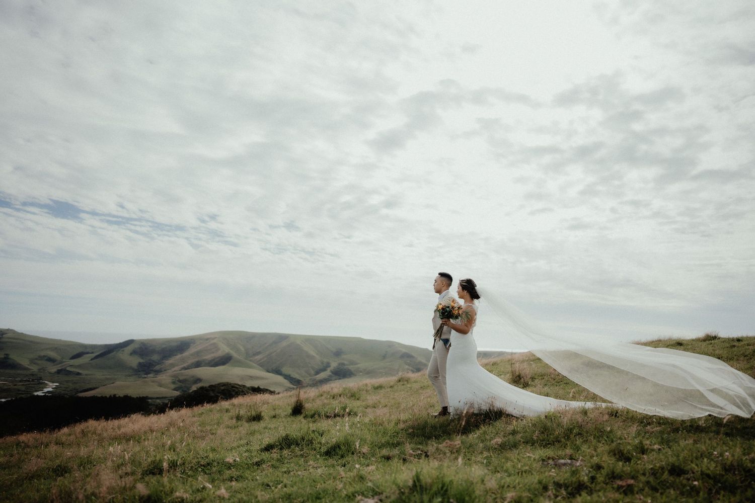 Bride and groom stand on a windswept hilltop as her long veil blows across a moody cloudy sky.