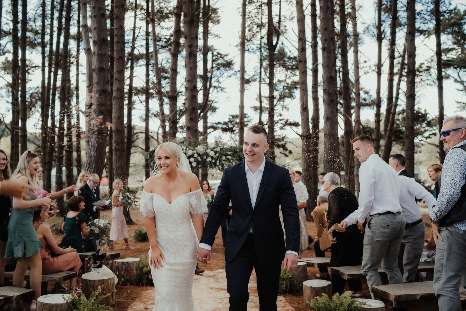 Smiling bride in a lace gown walks hand in hand with her groom down a pine forest aisle past seated wedding guests.