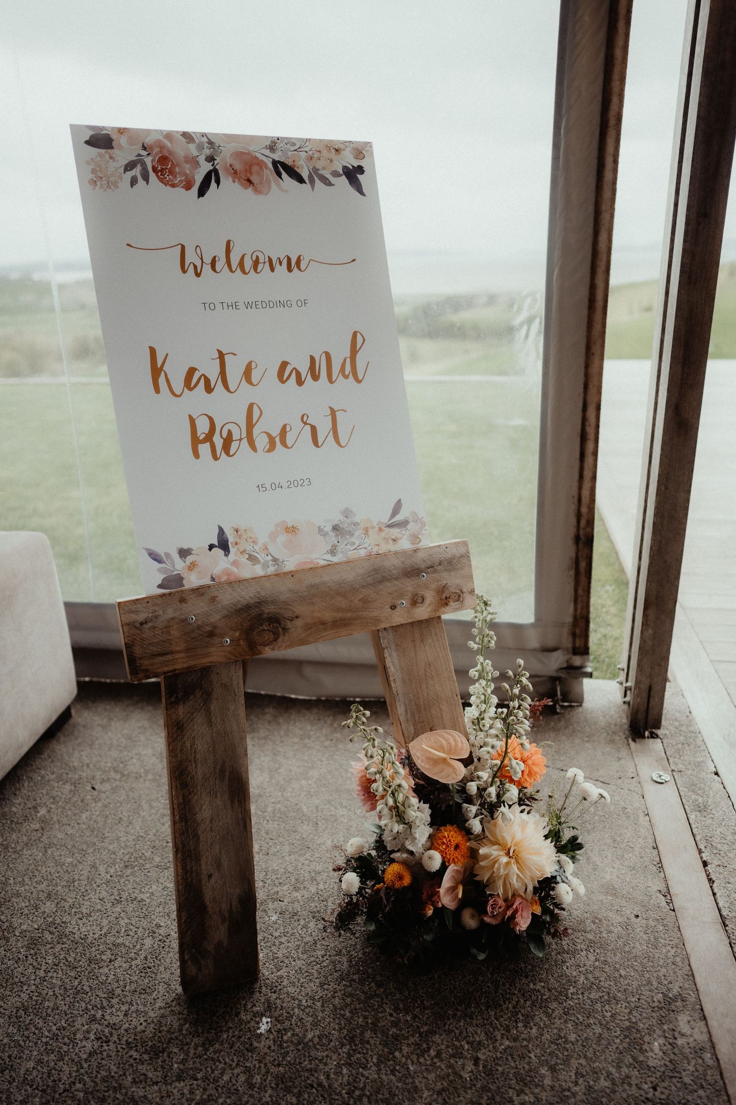 Wooden easel holds a floral welcome sign for Kate and Robert's wedding, beside an orange and white flower arrangement.